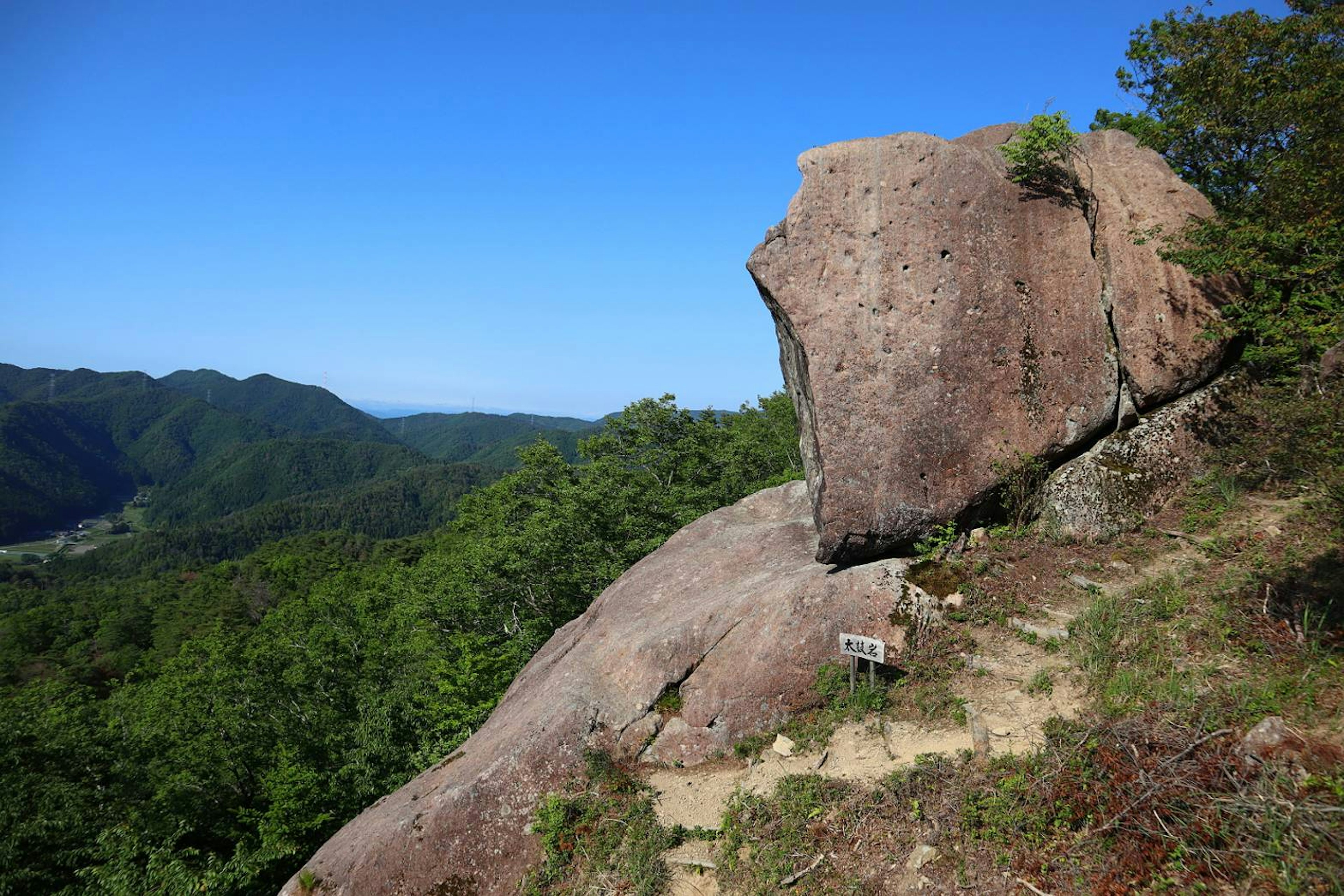 Large rock formation against a clear blue sky surrounded by green trees and hills