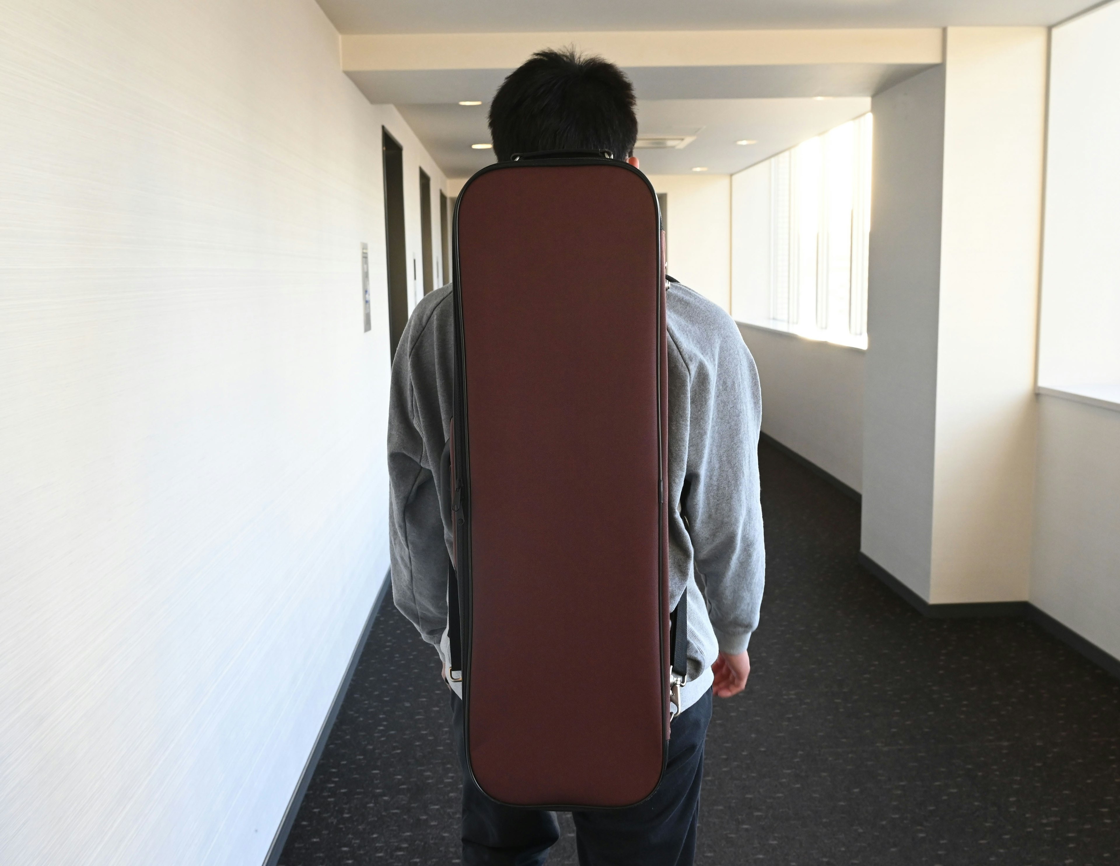 Man walking in a hallway with a brown guitar case on his back