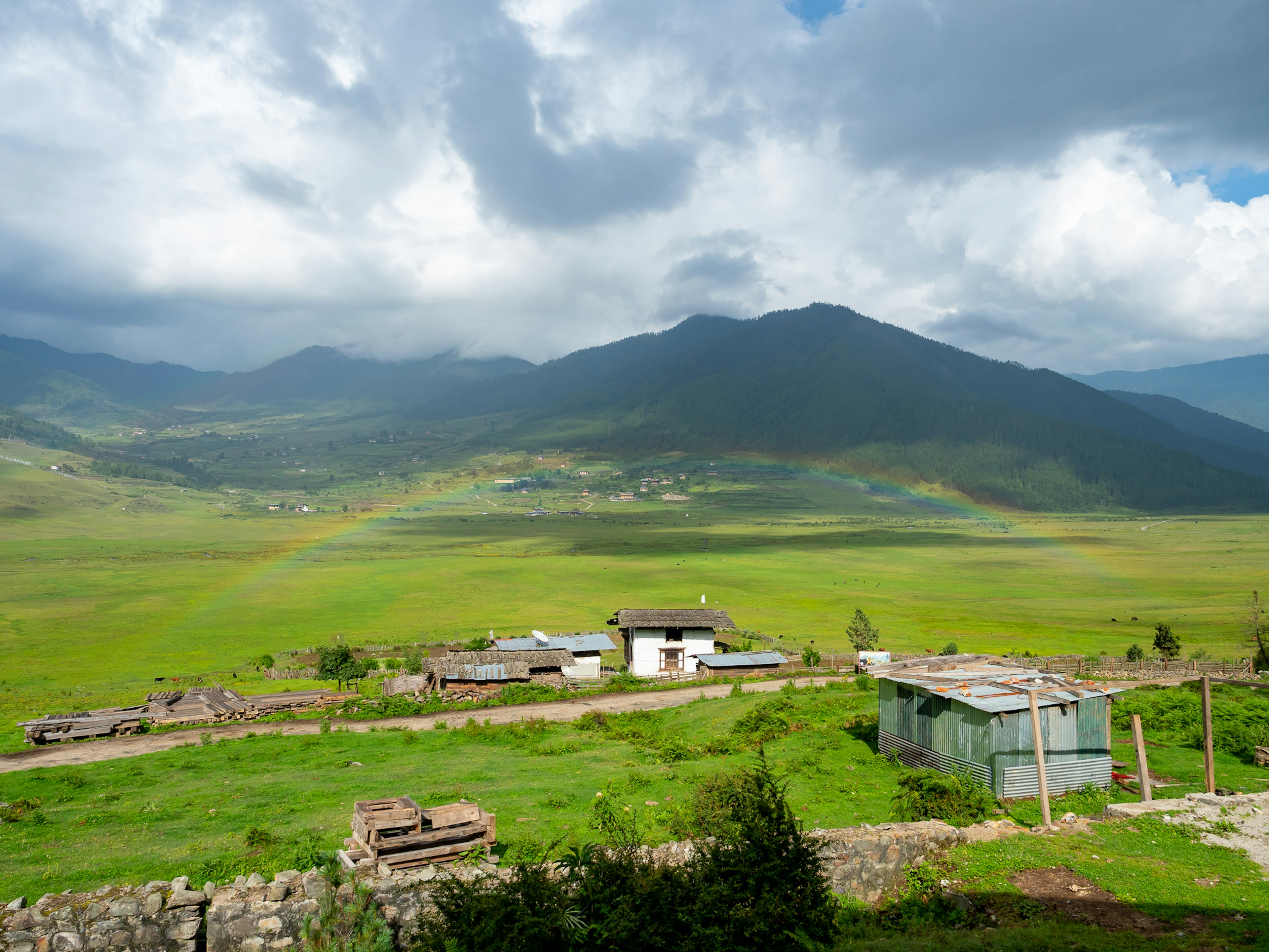 Vista escénica de montañas y campos verdes bajo un cielo nublado con un toque de arco iris