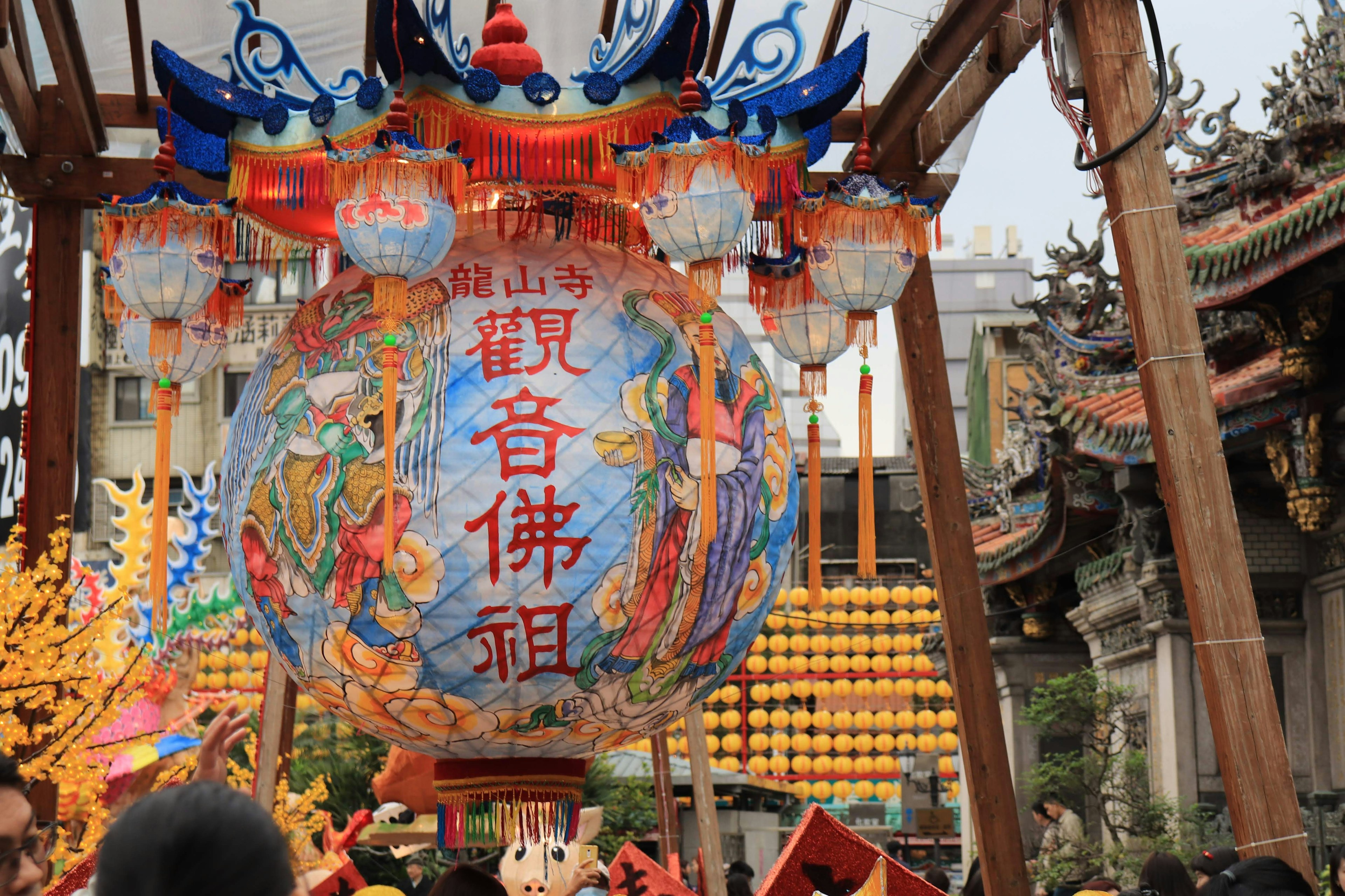 Scène de festival vibrante avec des lanternes colorées incluant une grande lanterne décorée de caractères chinois et de figures mythologiques