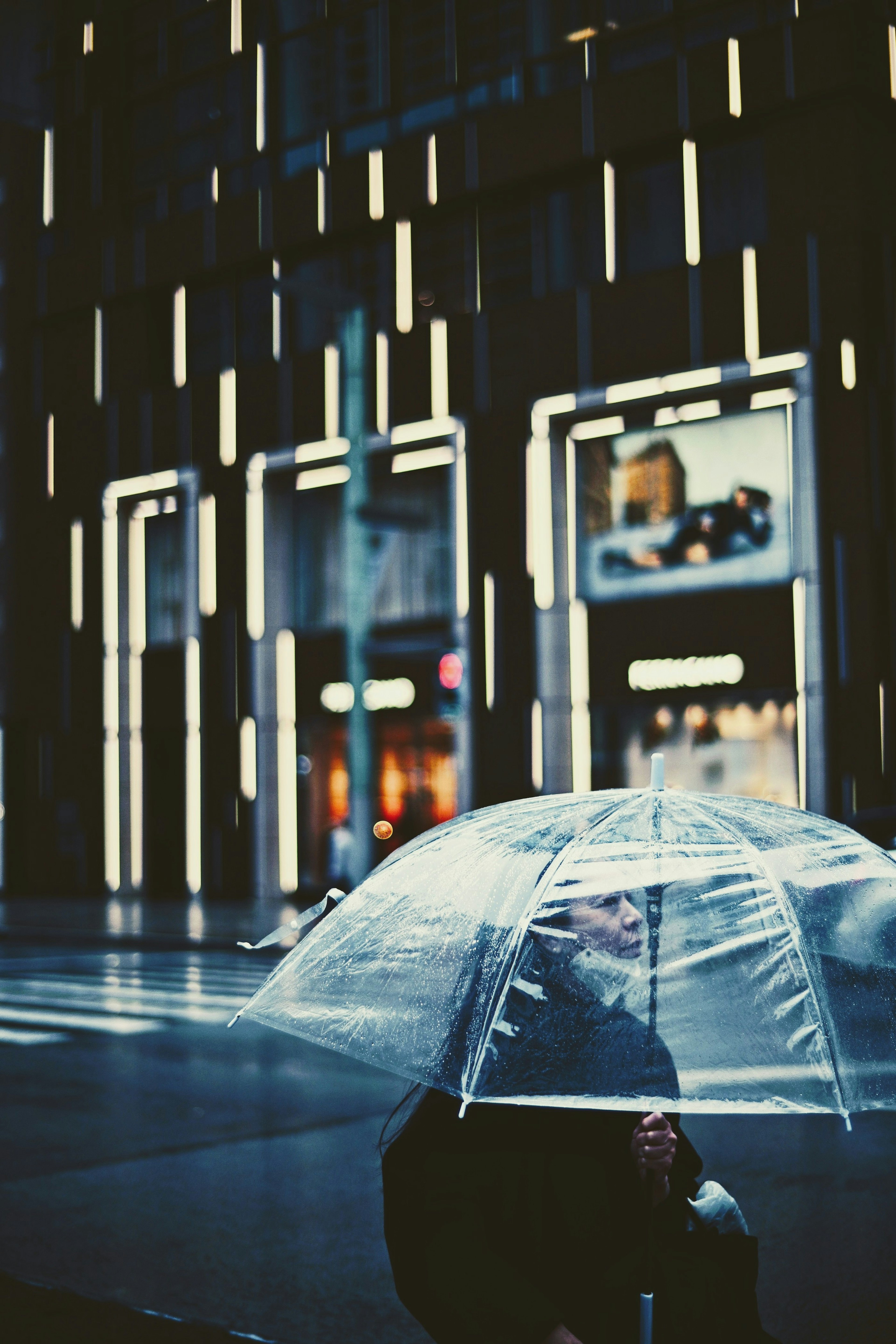 Person holding a transparent umbrella in front of brightly lit buildings on a rainy street