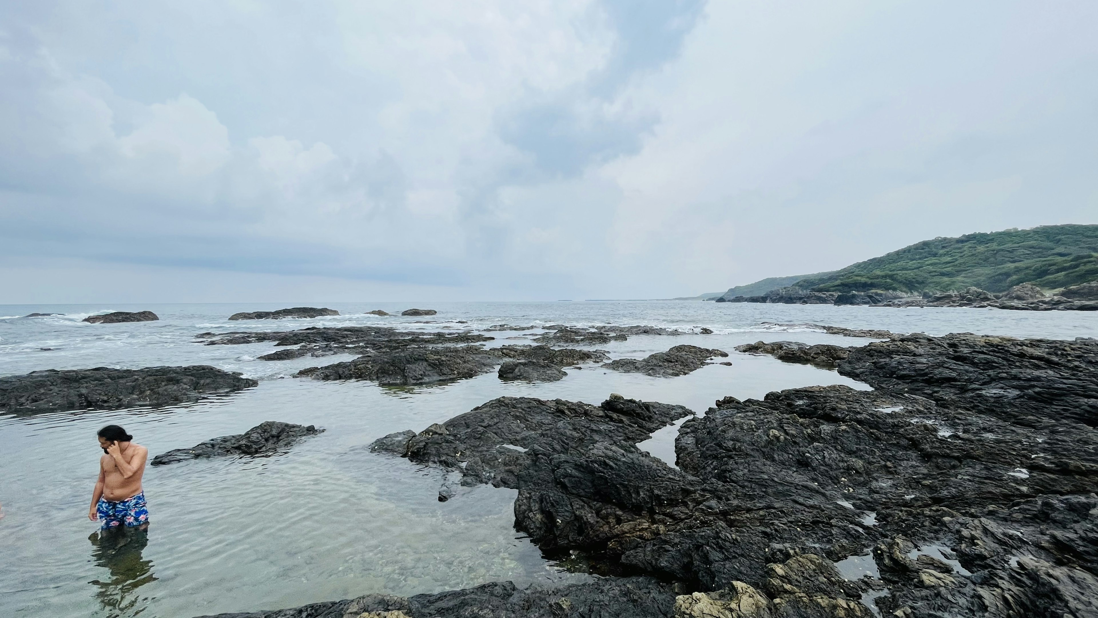 Seorang anak bermain di pantai berbatu dengan laut tenang dan langit mendung