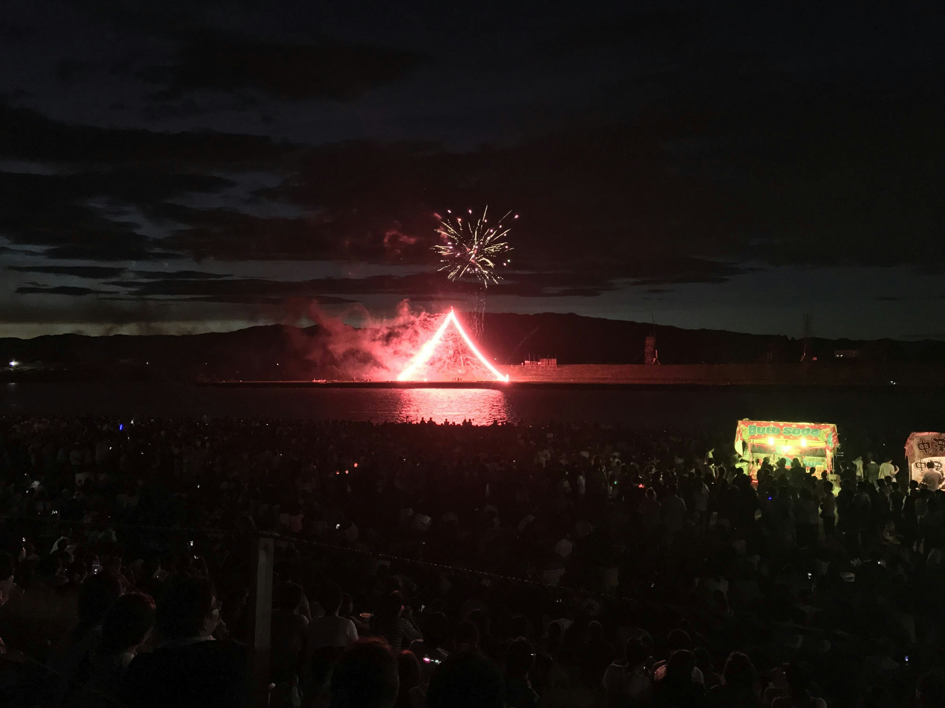 Escena de festival con fuegos artificiales formando una figura triangular roja en el cielo nocturno