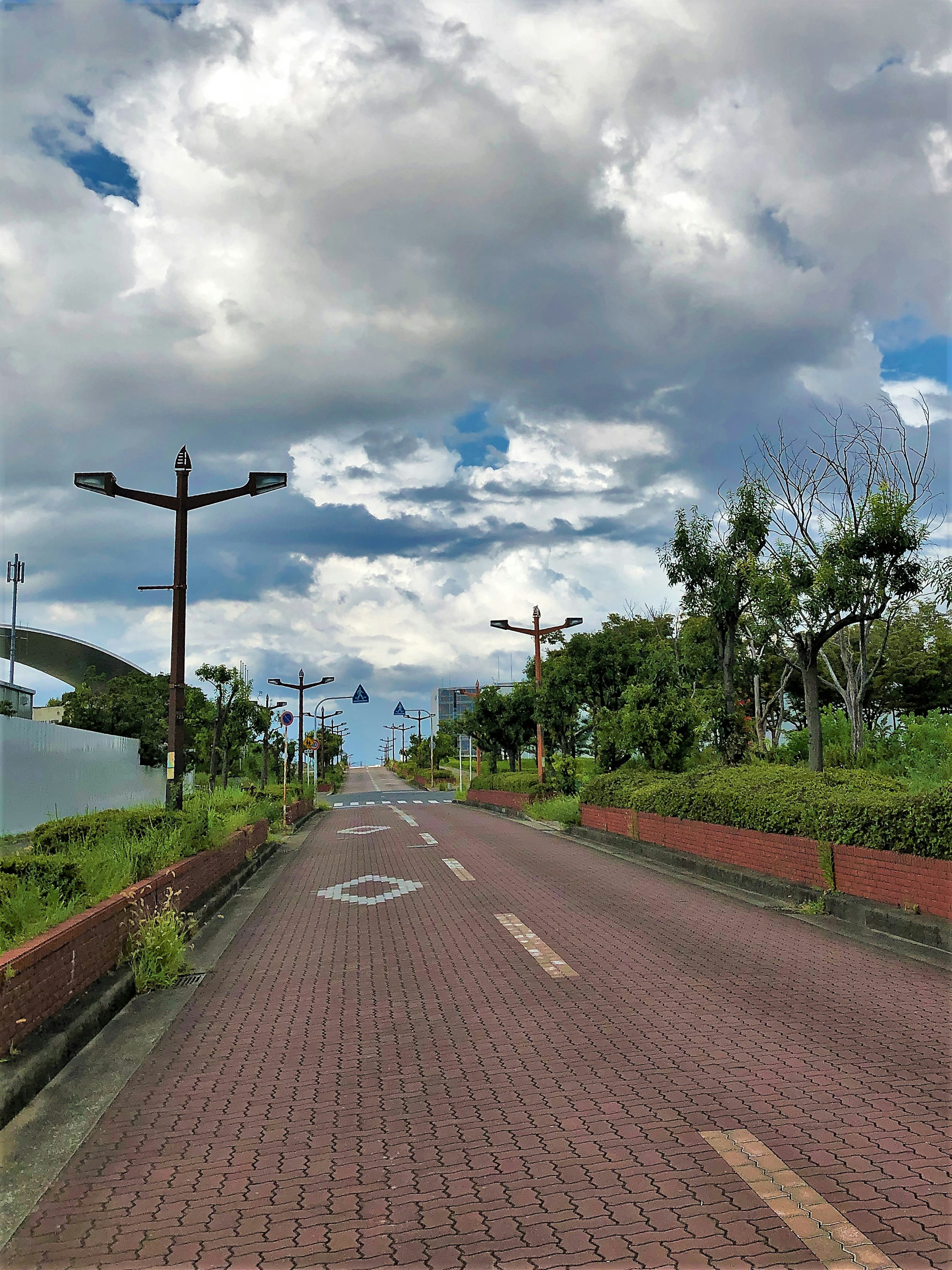 Jalan yang dipaving dikelilingi pepohonan di bawah langit biru dengan awan