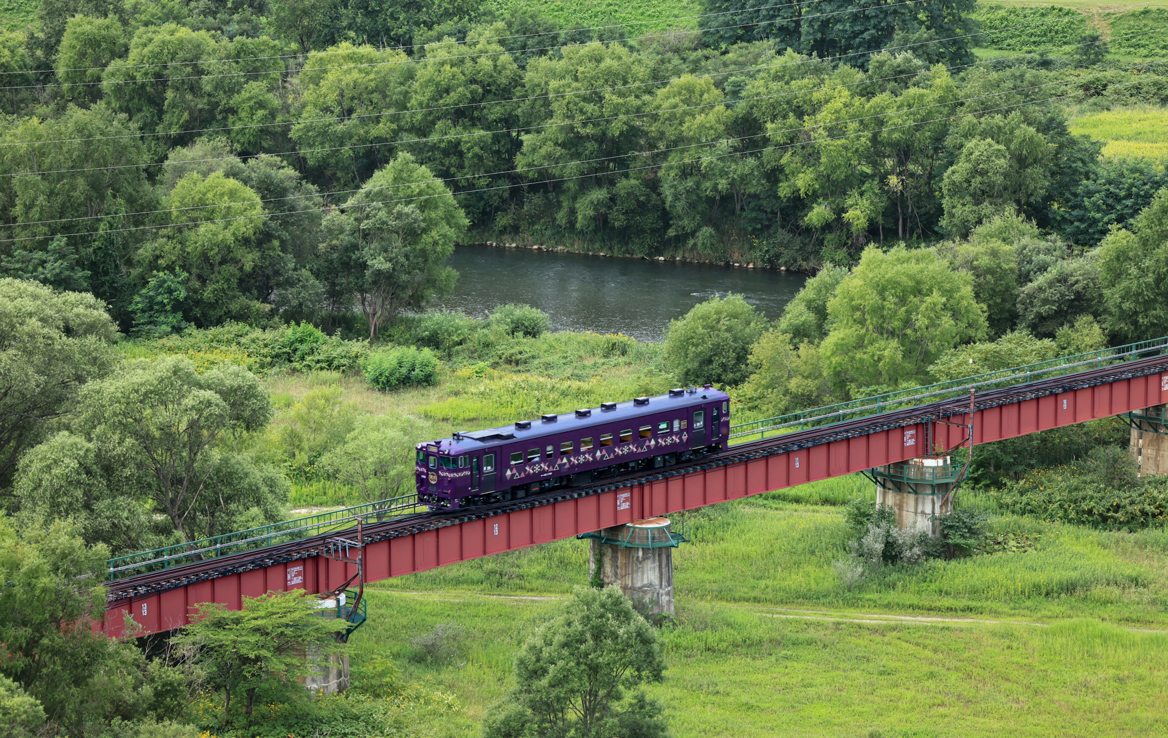 Sebuah kereta ungu melintasi jembatan merah dikelilingi pemandangan hijau subur