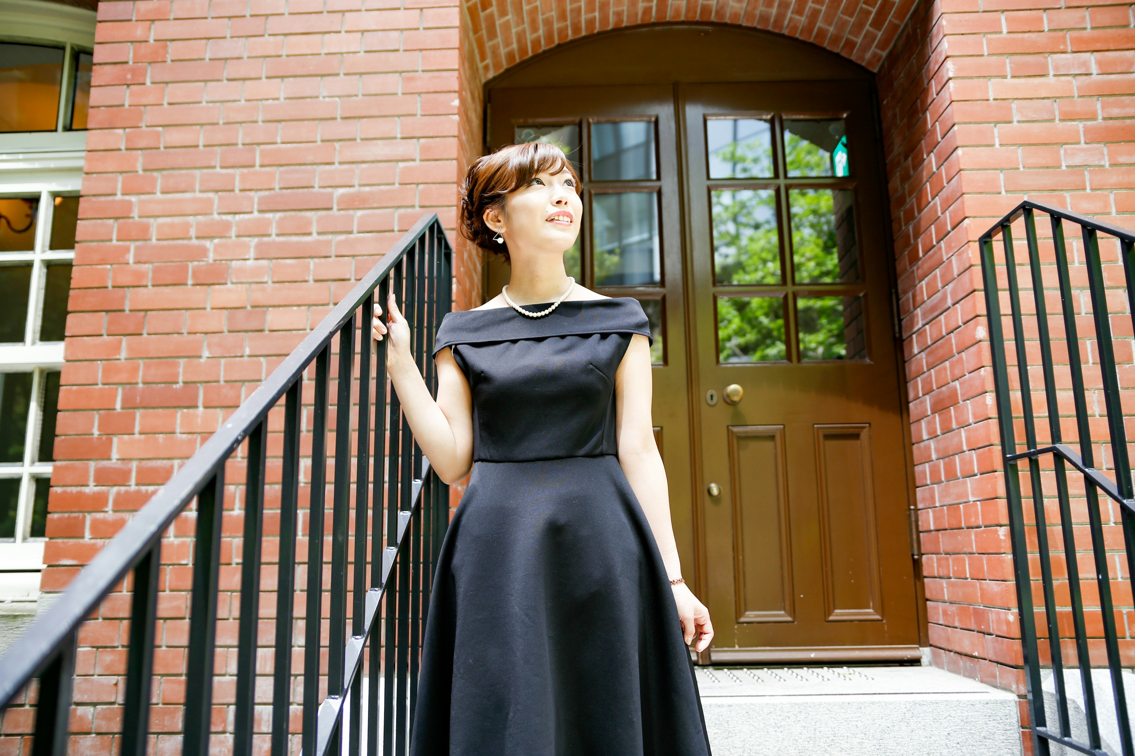 A woman in a black dress standing on stairs with a brick building and wooden door in the background