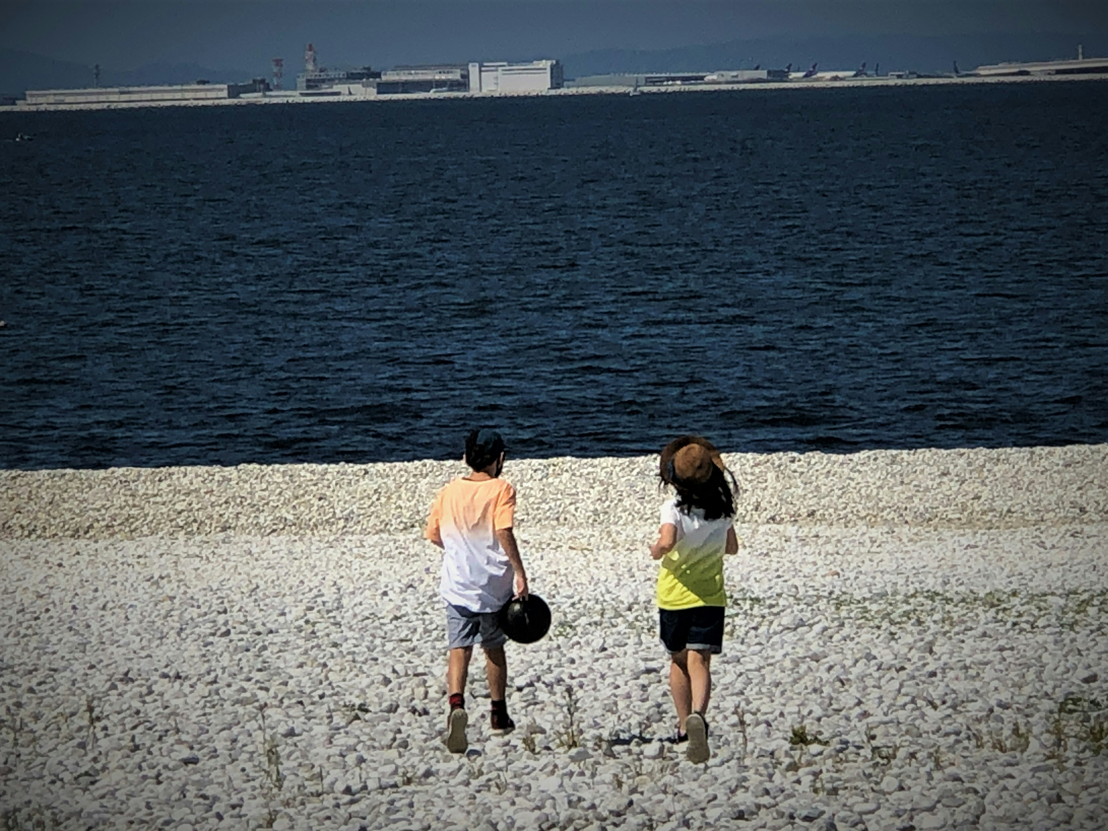 二人が砂利のビーチを歩いている後ろ姿の写真 海の青と空の色が美しい