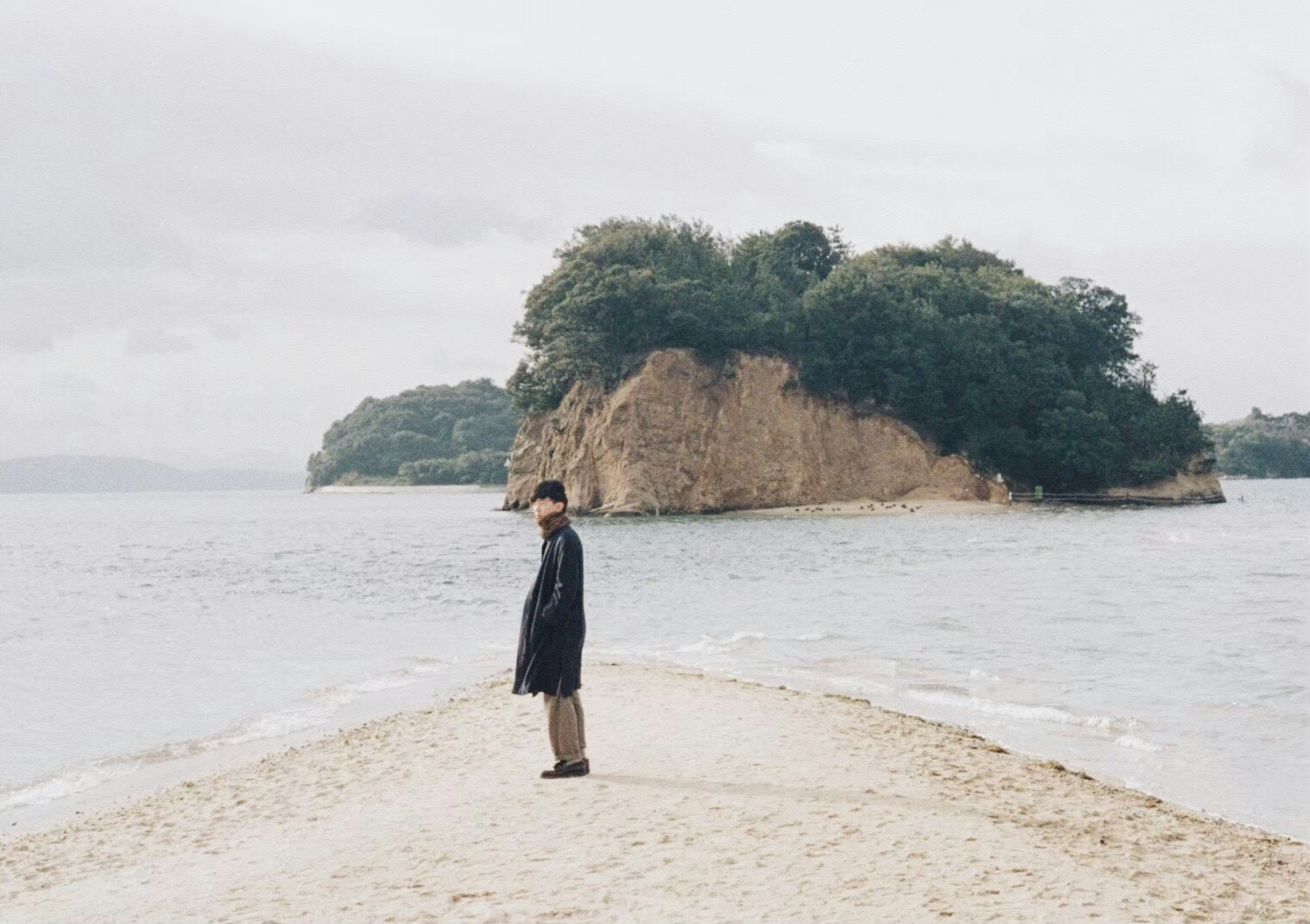 A man standing on a sandy path by the sea with an island in the background