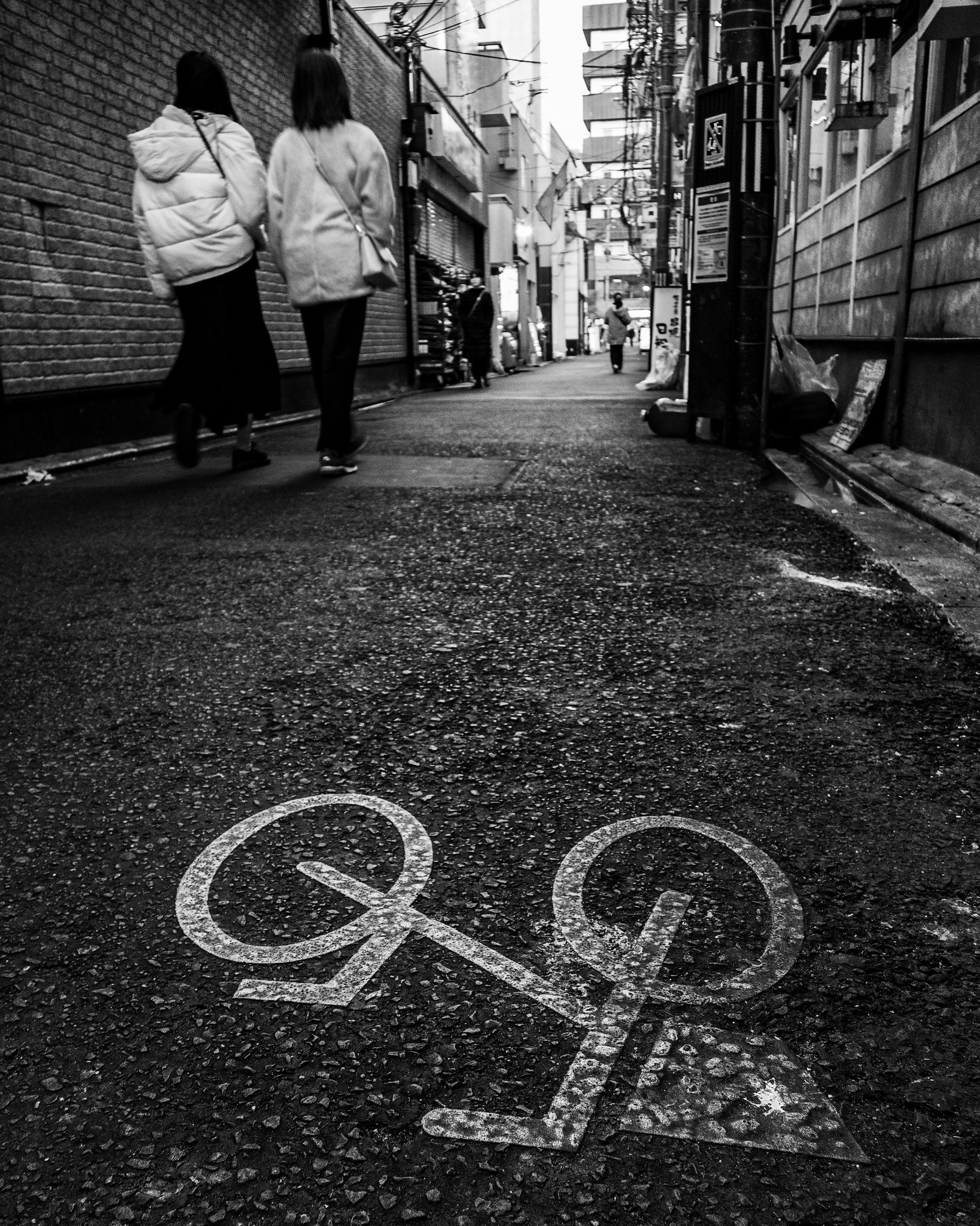 Two women walking down a narrow alley with a bicycle symbol on the ground