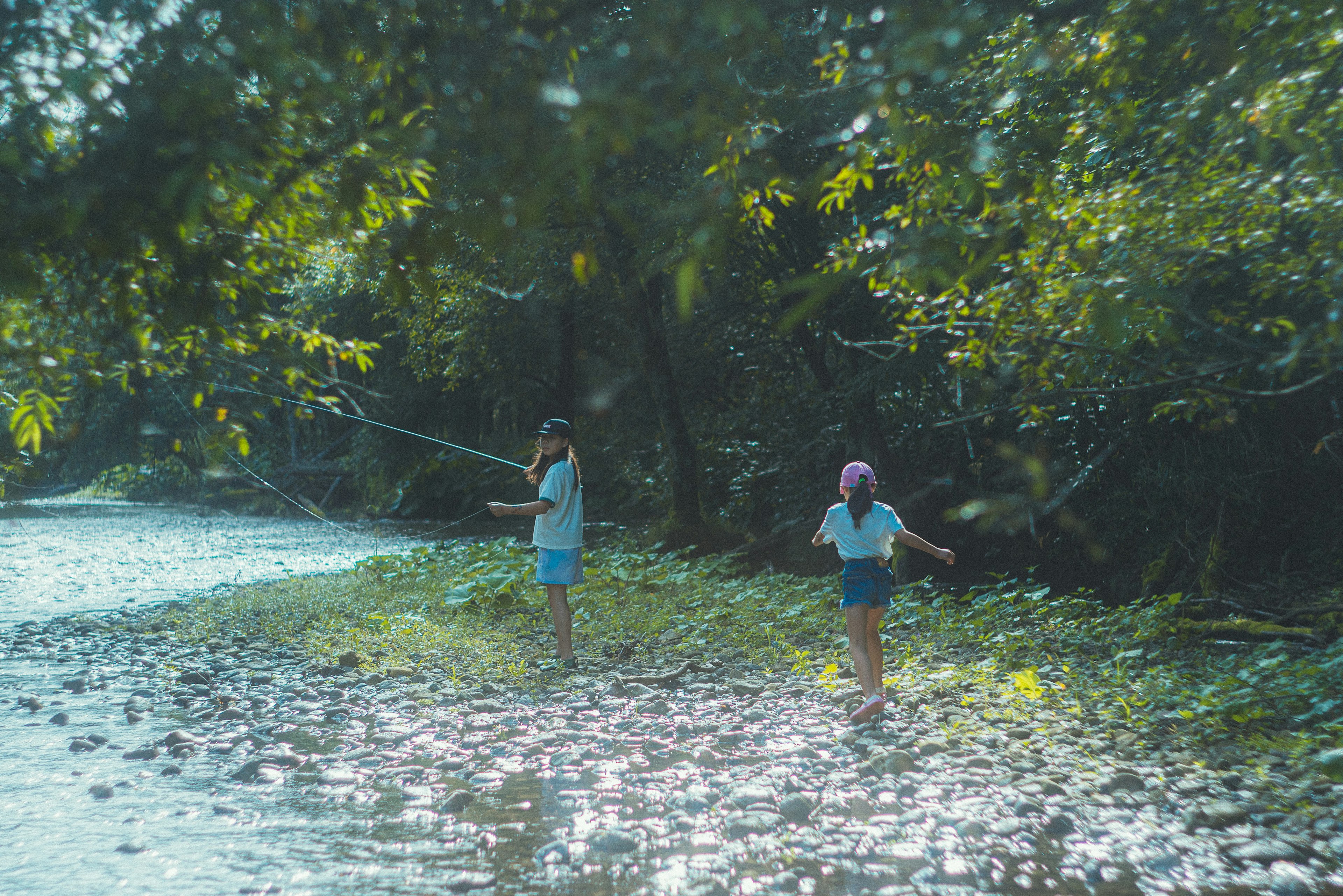 Dua anak memancing di tepi sungai dikelilingi oleh pepohonan hijau