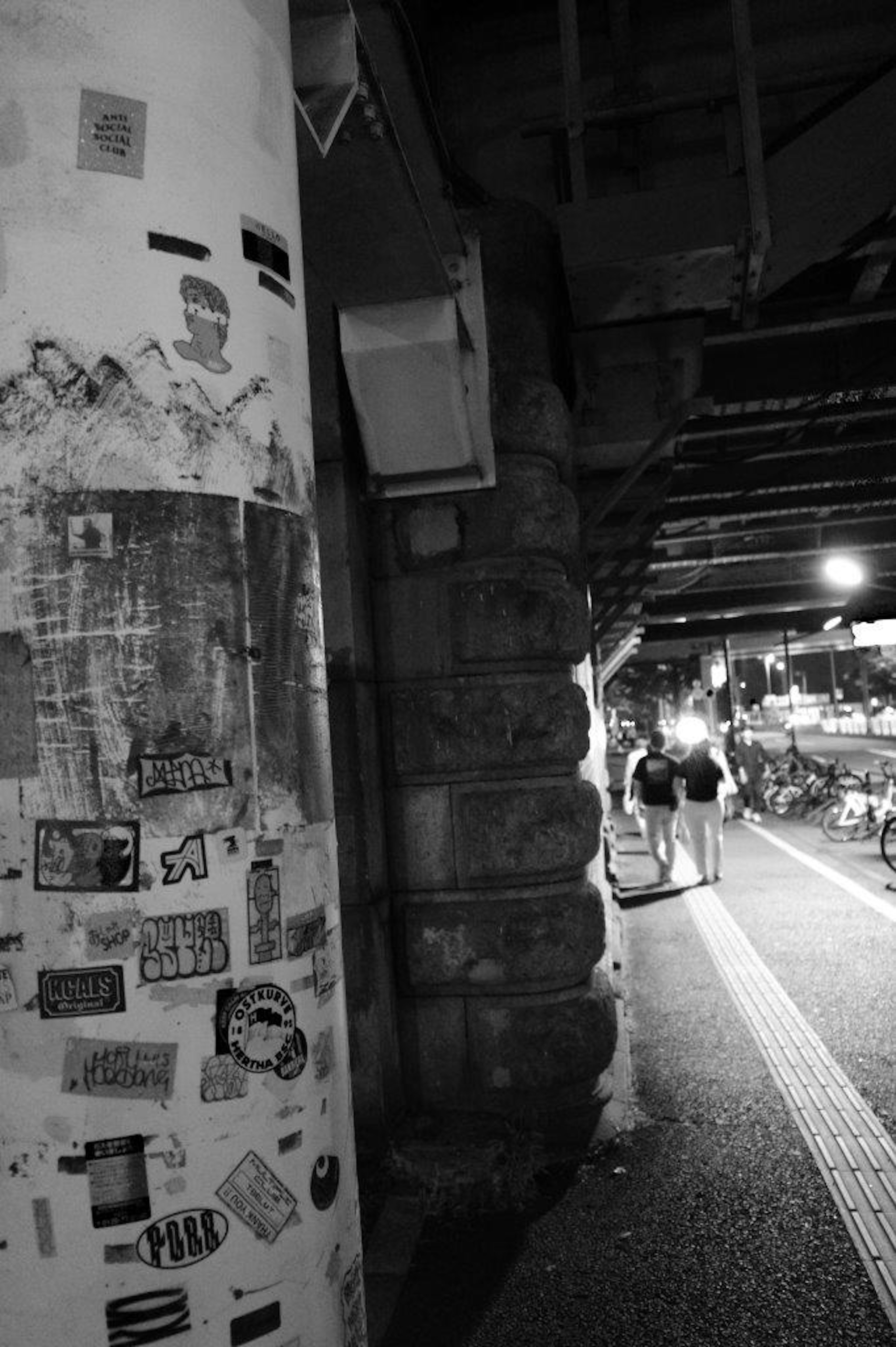 Escena urbana en blanco y negro con un pilar cubierto de carteles y una acera