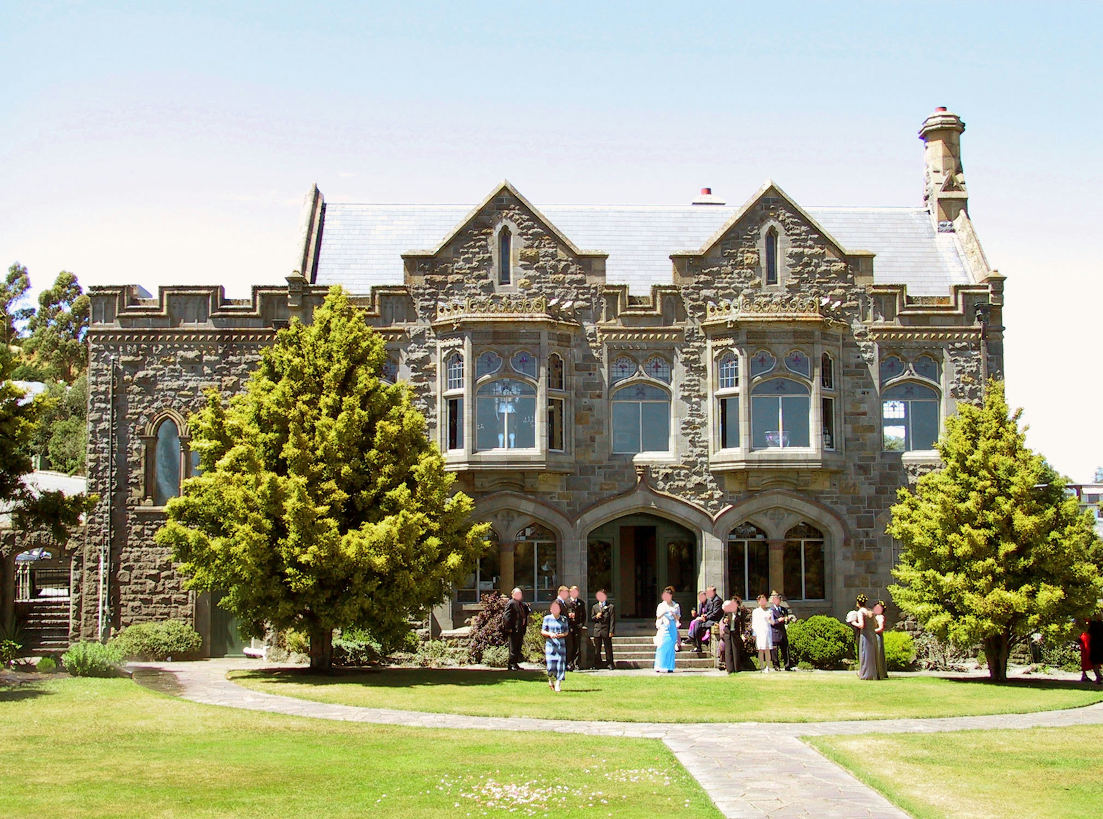 People gathered in front of a beautiful stone house surrounded by green trees