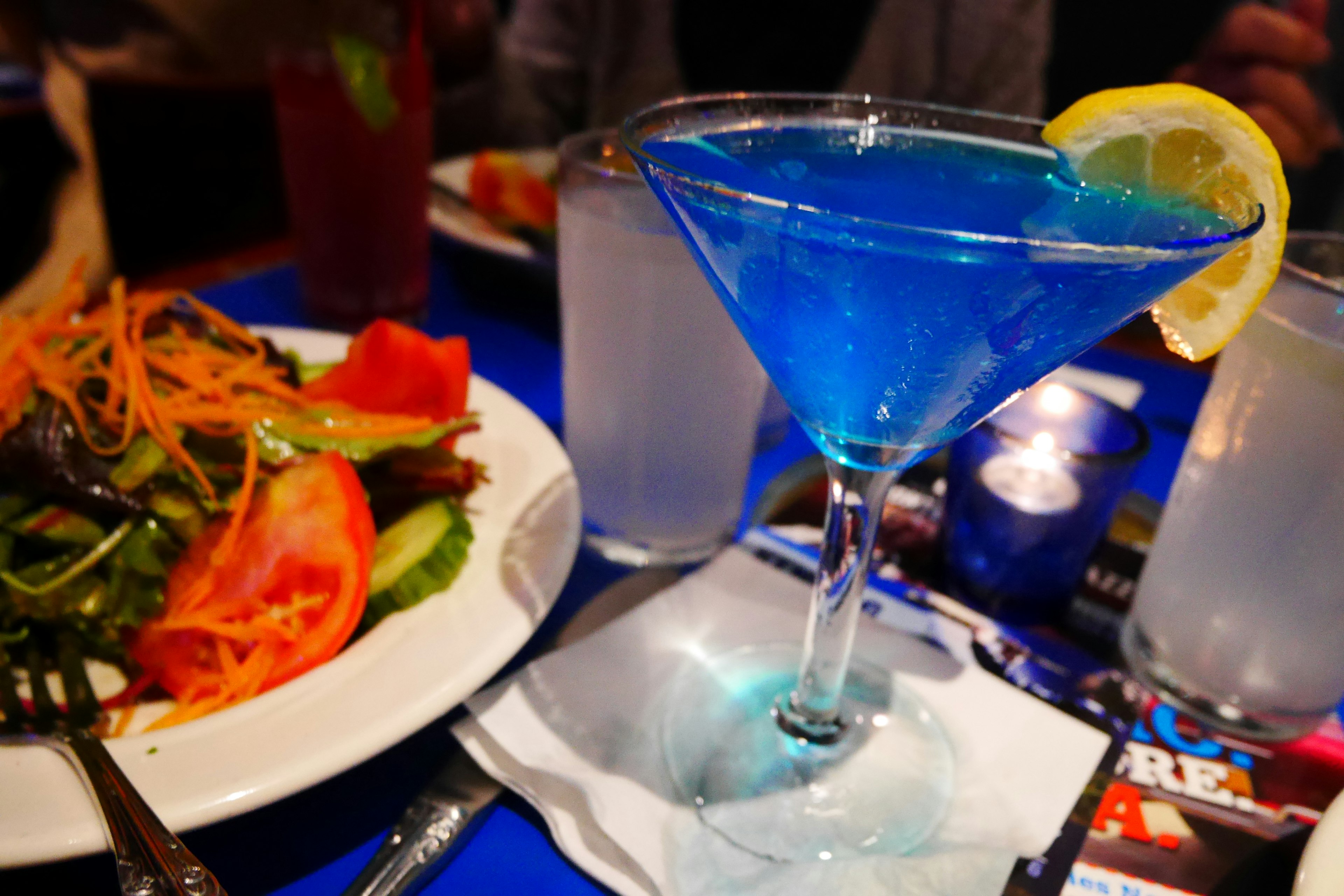 Ein blauer Cocktail mit einer Zitronenscheibe neben einem frischen Salat auf einem Tisch