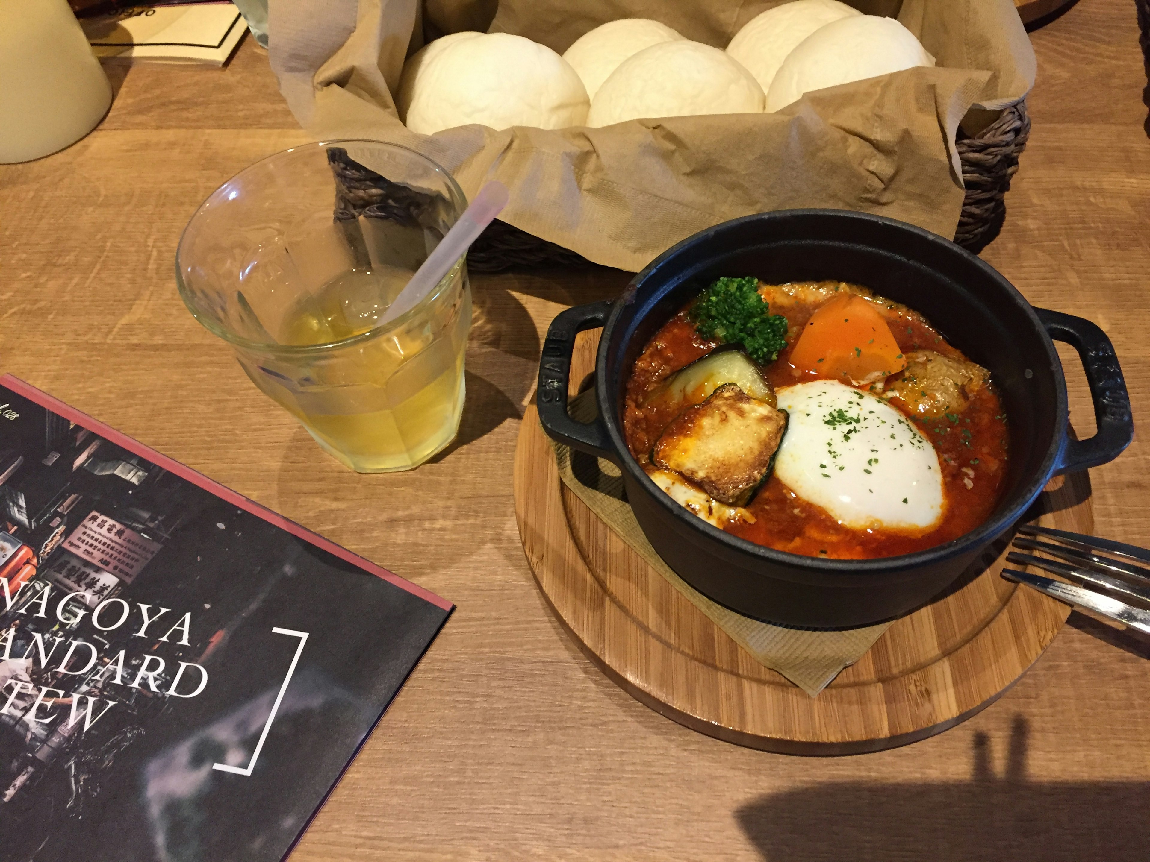 Una olla negra llena de guiso con verduras y un huevo al lado de una canasta de pan en una mesa de madera