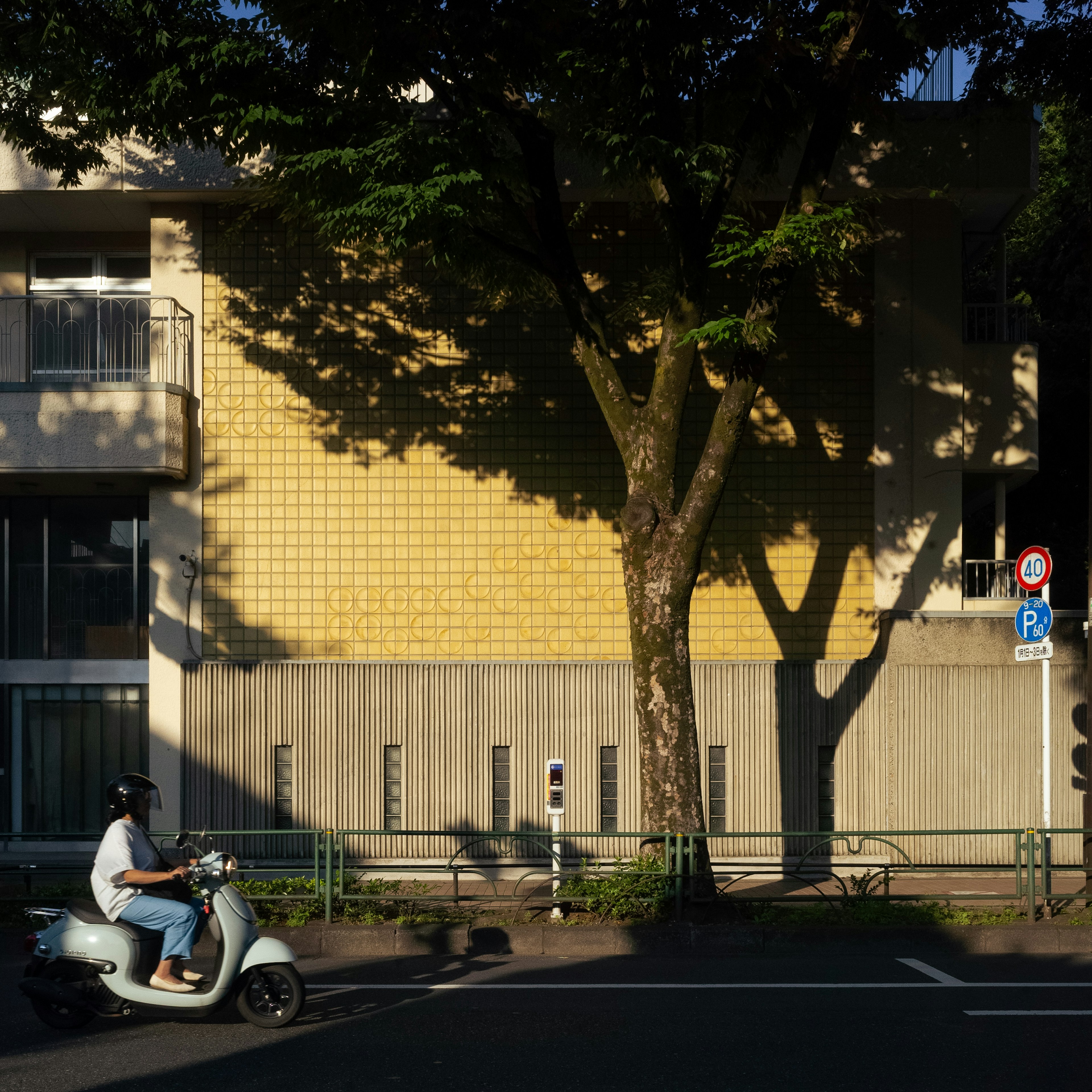 黄色いタイルの壁のある建物と緑の木の影のある風景でスクーターに乗る人