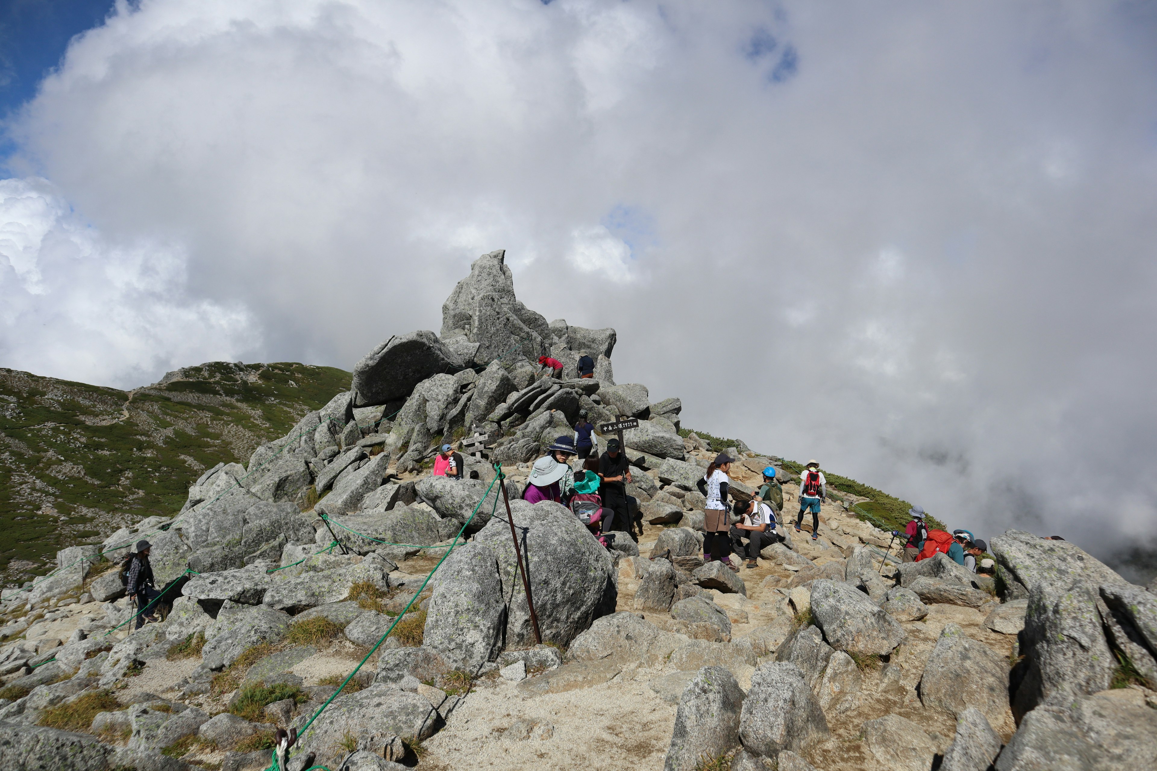 Personas haciendo senderismo en la cima de una montaña rocosa con nubes al fondo