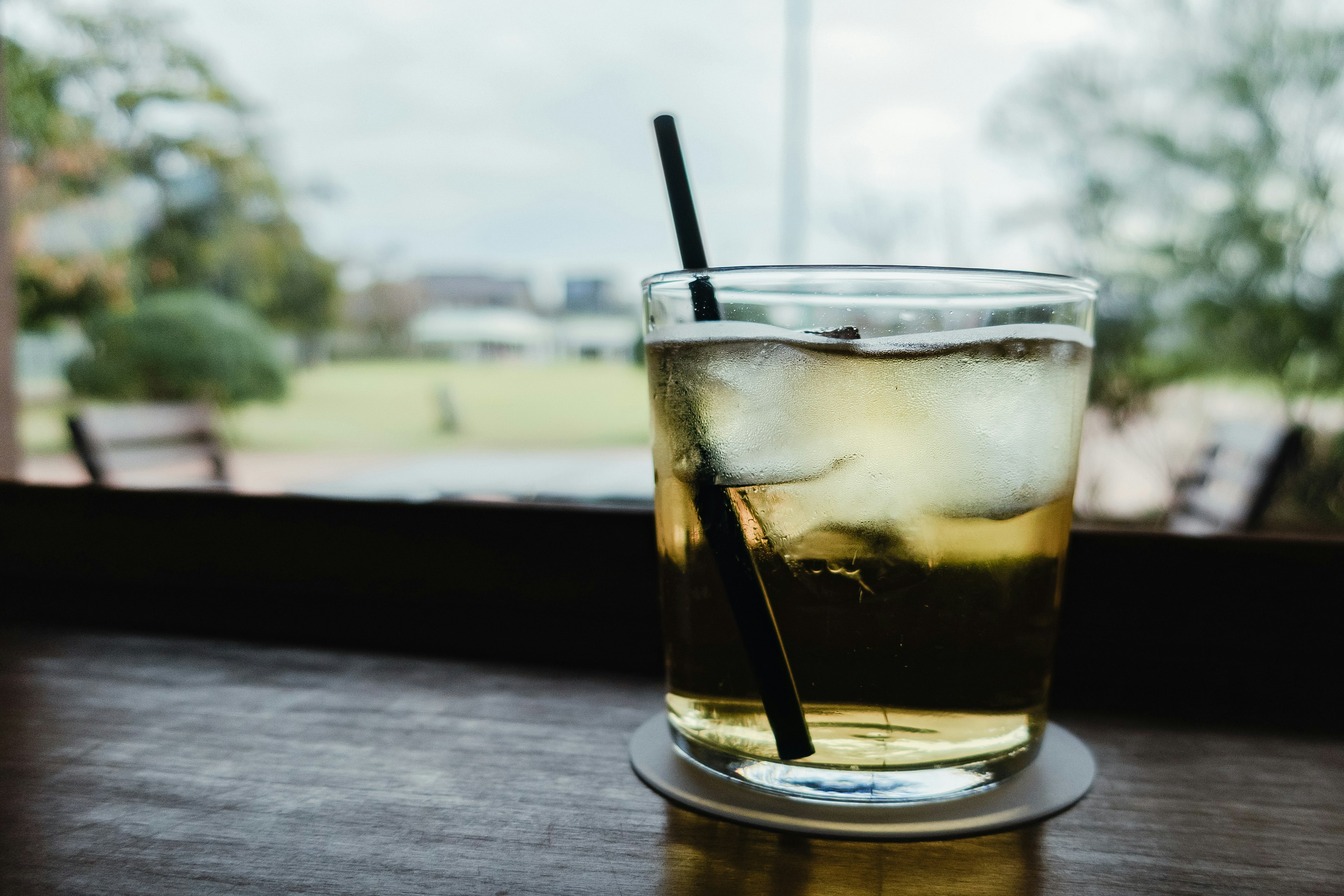 A cocktail glass with ice sits on a windowsill with a park view in the background