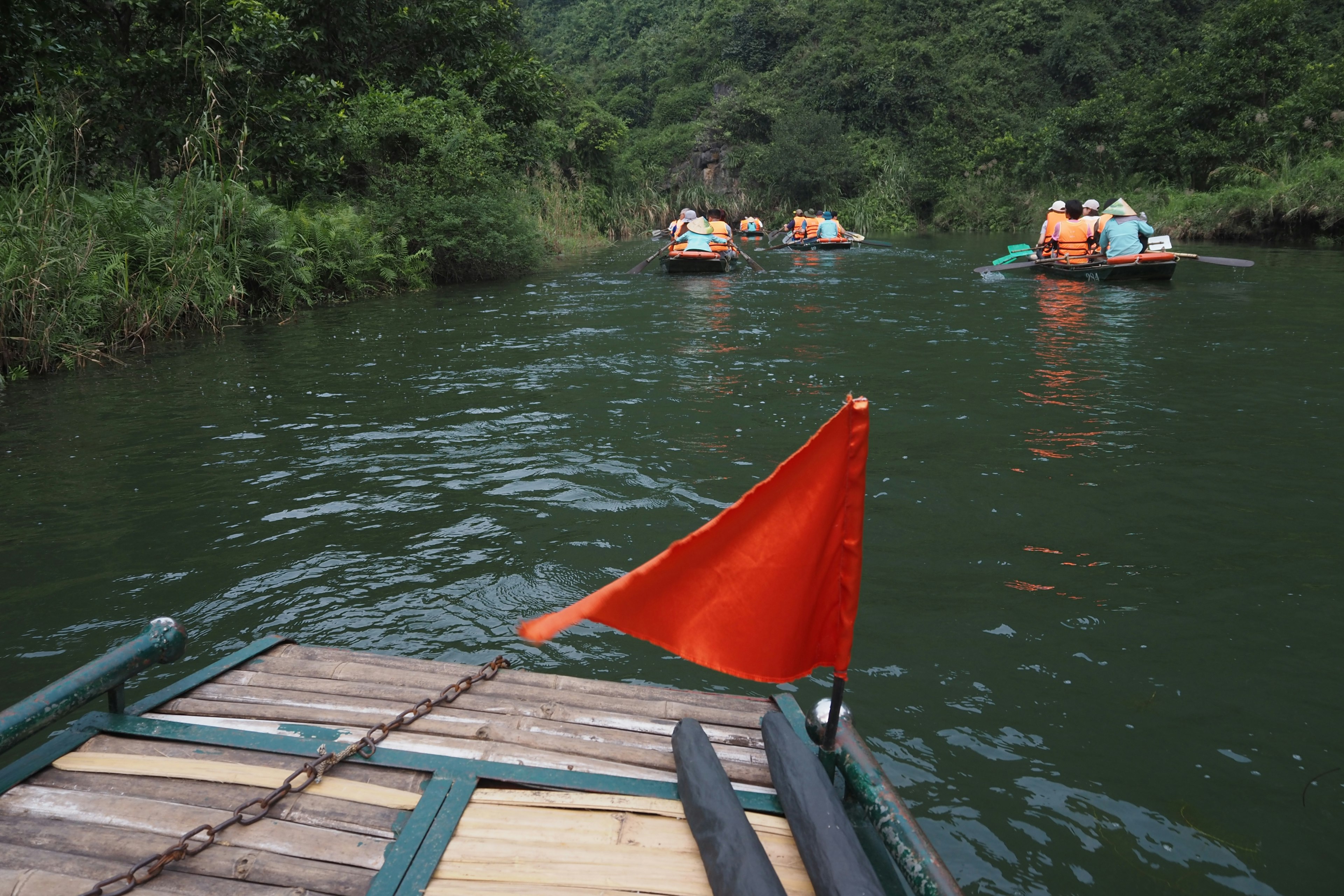 Kleine Boote auf einem grünen Fluss, umgeben von üppiger Vegetation, mit einer roten Flagge im Vordergrund