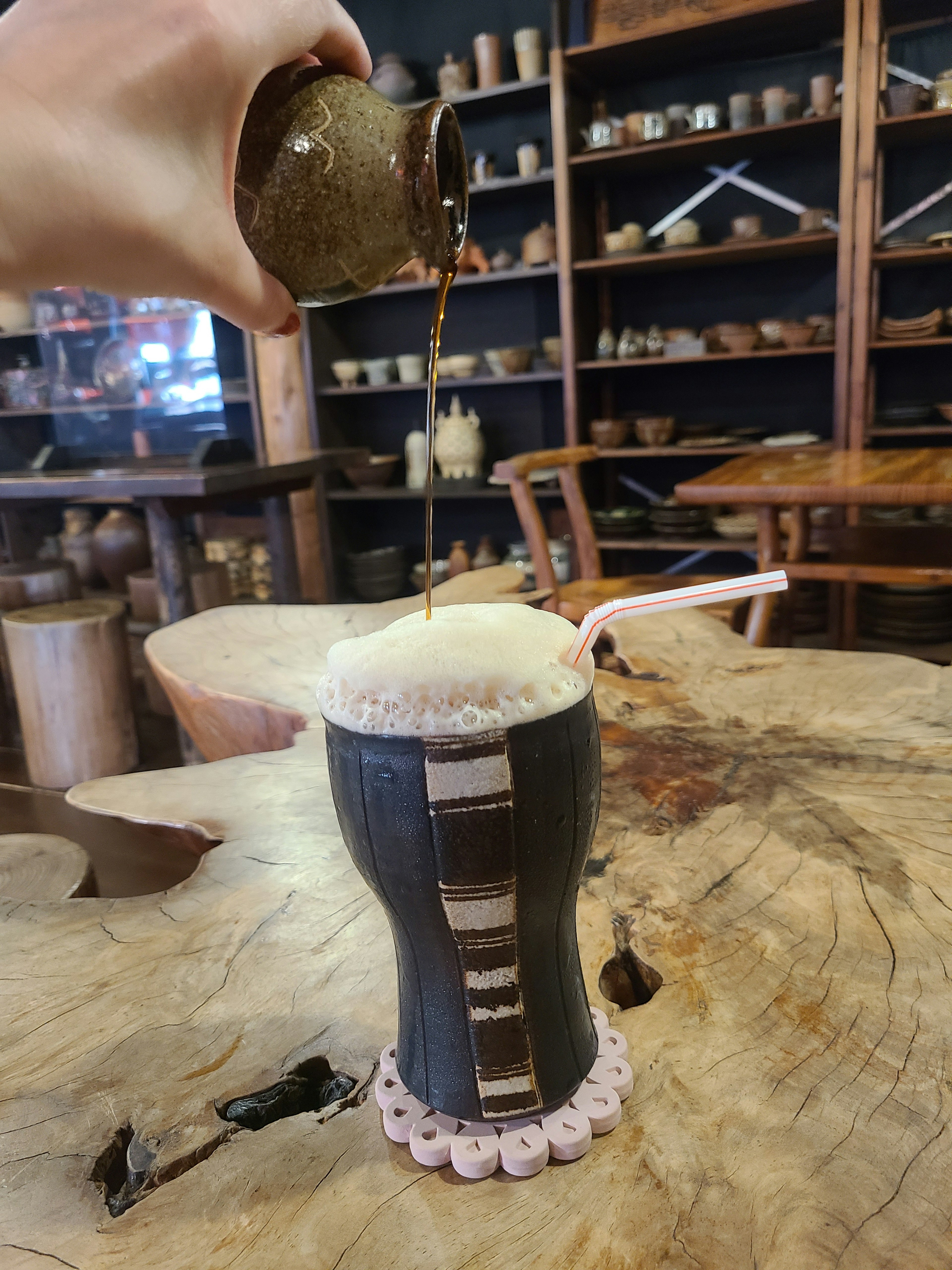 Une main versant une boisson dans une tasse noire avec de la mousse sur le dessus, posée sur une table en bois