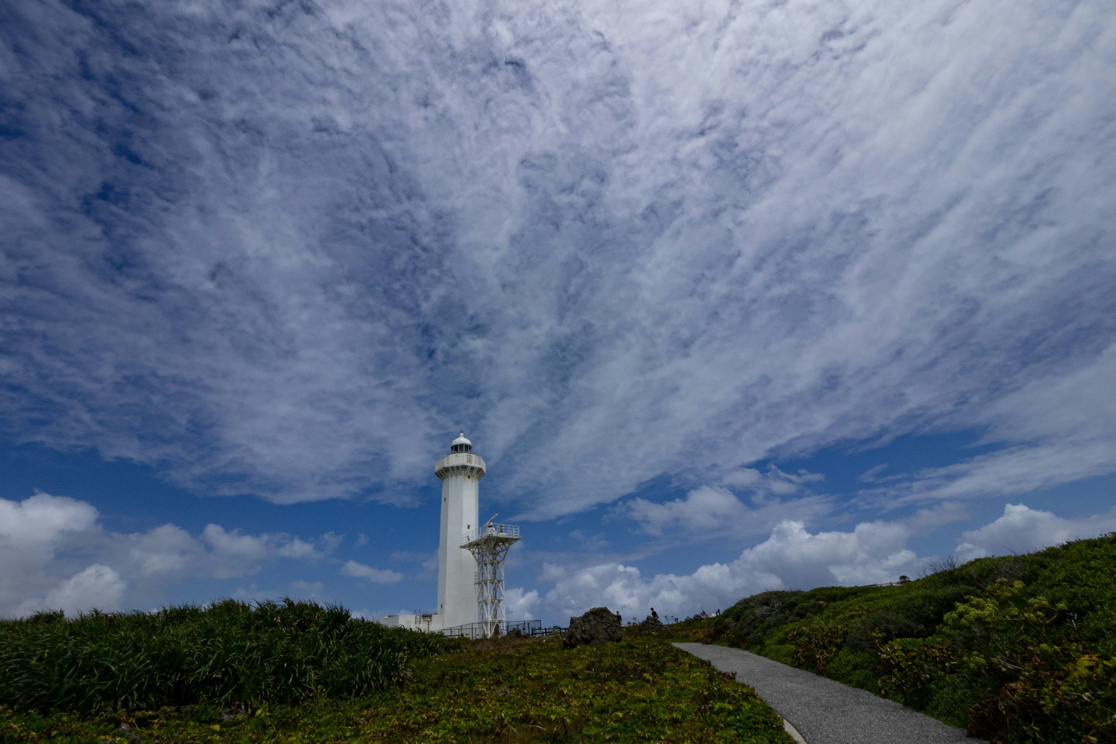 A white lighthouse standing under a blue sky with clouds and green hills