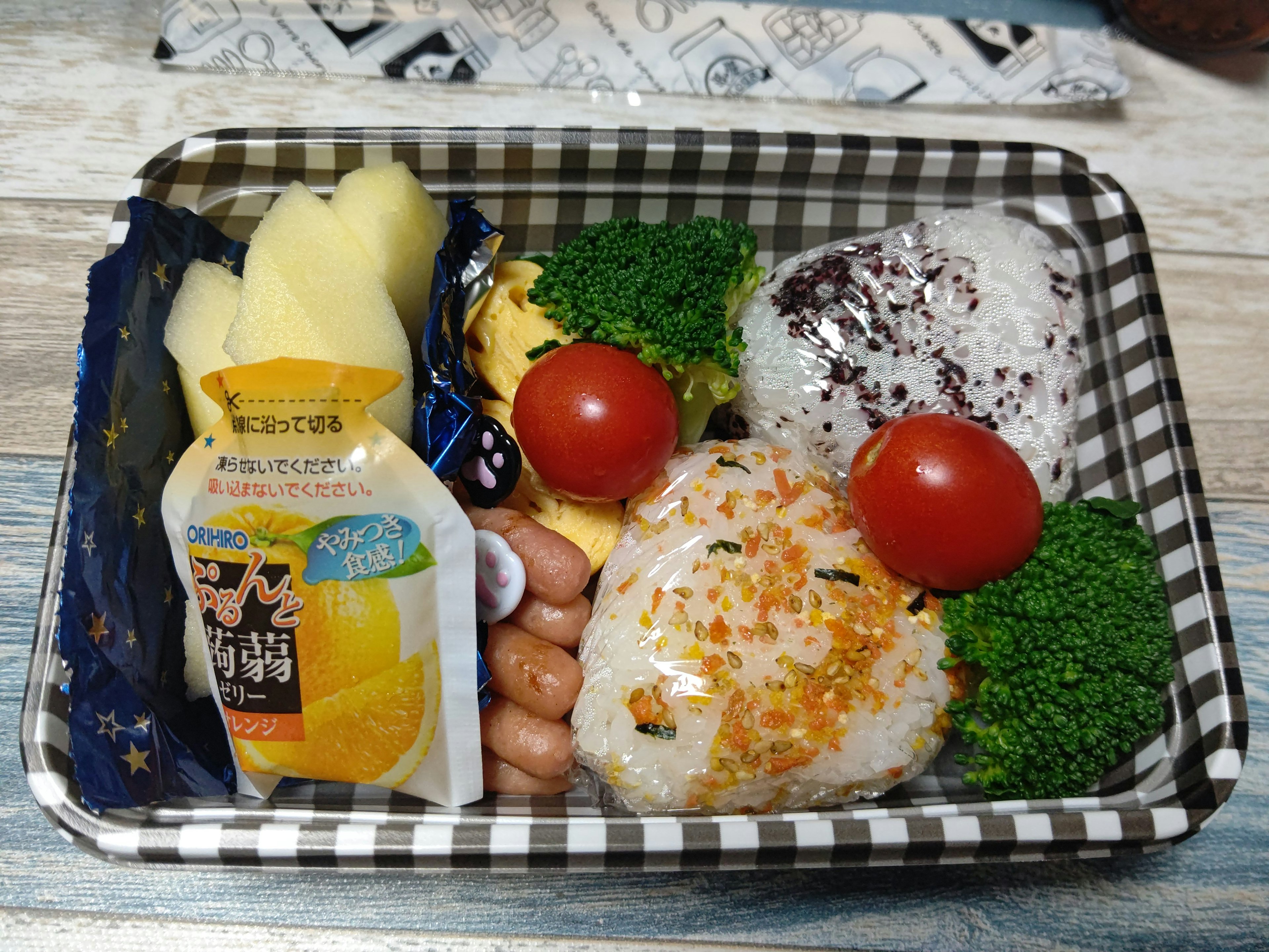 A bento box featuring rice balls, broccoli, cherry tomatoes, sausages, bananas, and a drink