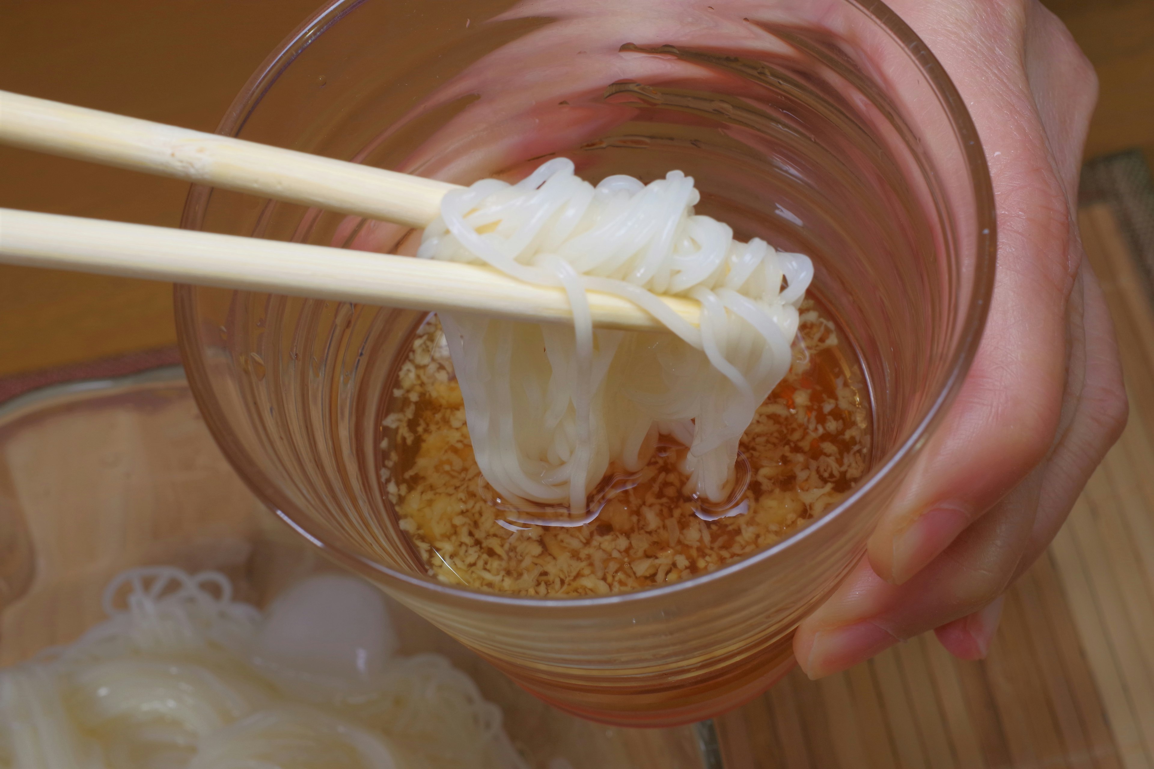 A hand holding chopsticks lifting cold noodles from a transparent glass