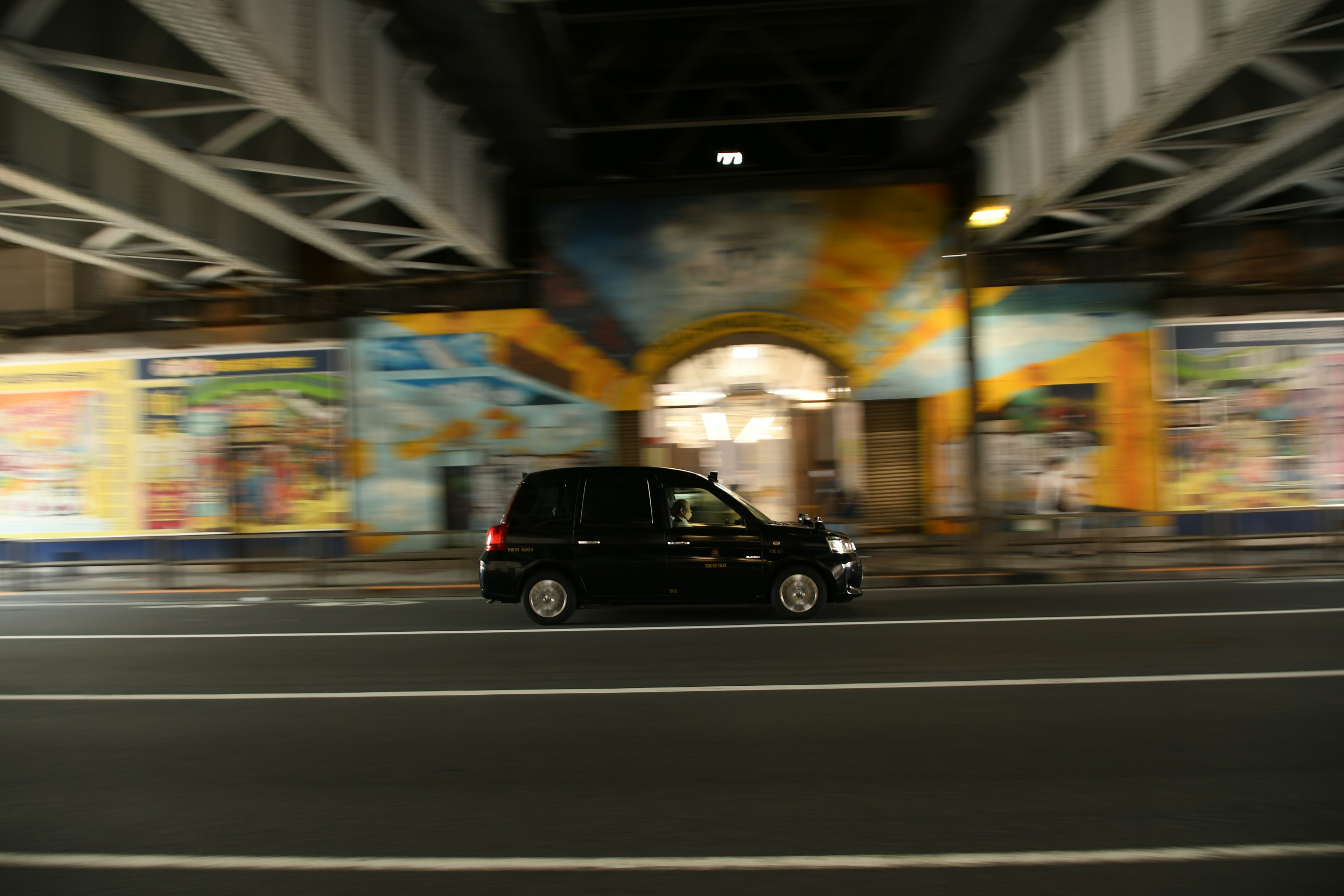 A black car driving under an overpass with colorful murals in the background