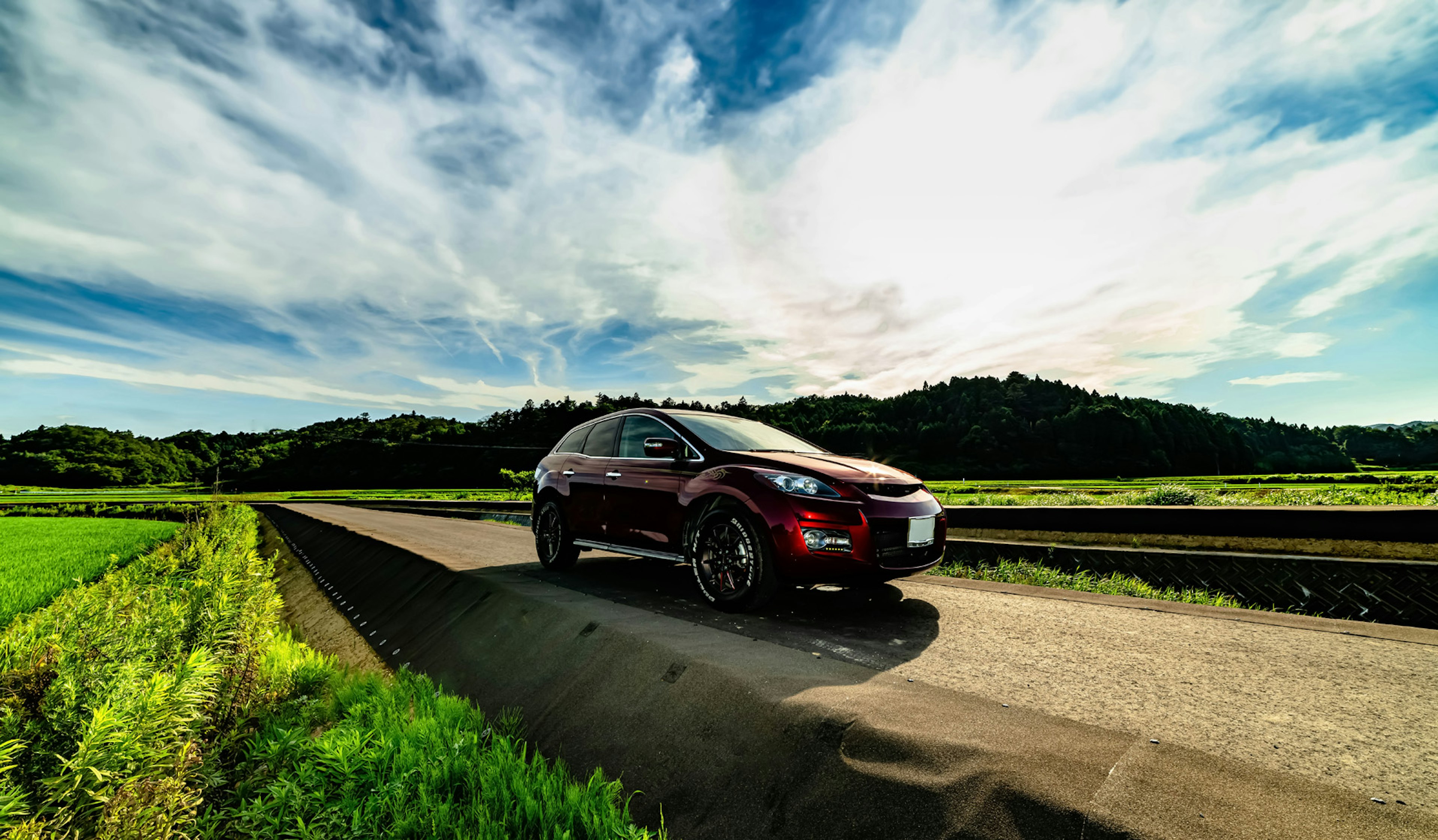 A red SUV driving on a rural road surrounded by green fields and blue sky