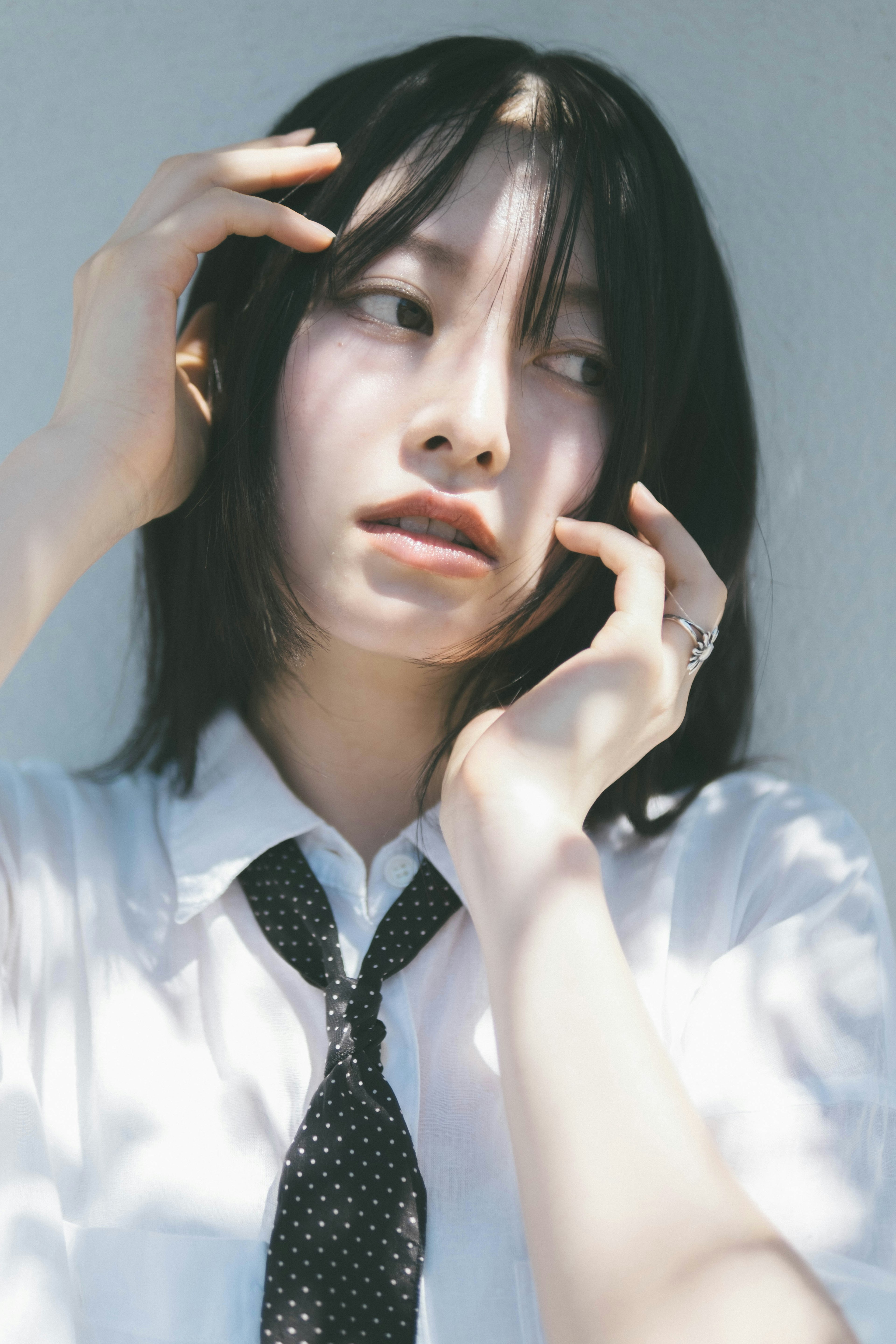 A woman with black hair wearing a white shirt and black tie touching her hair while deep in thought