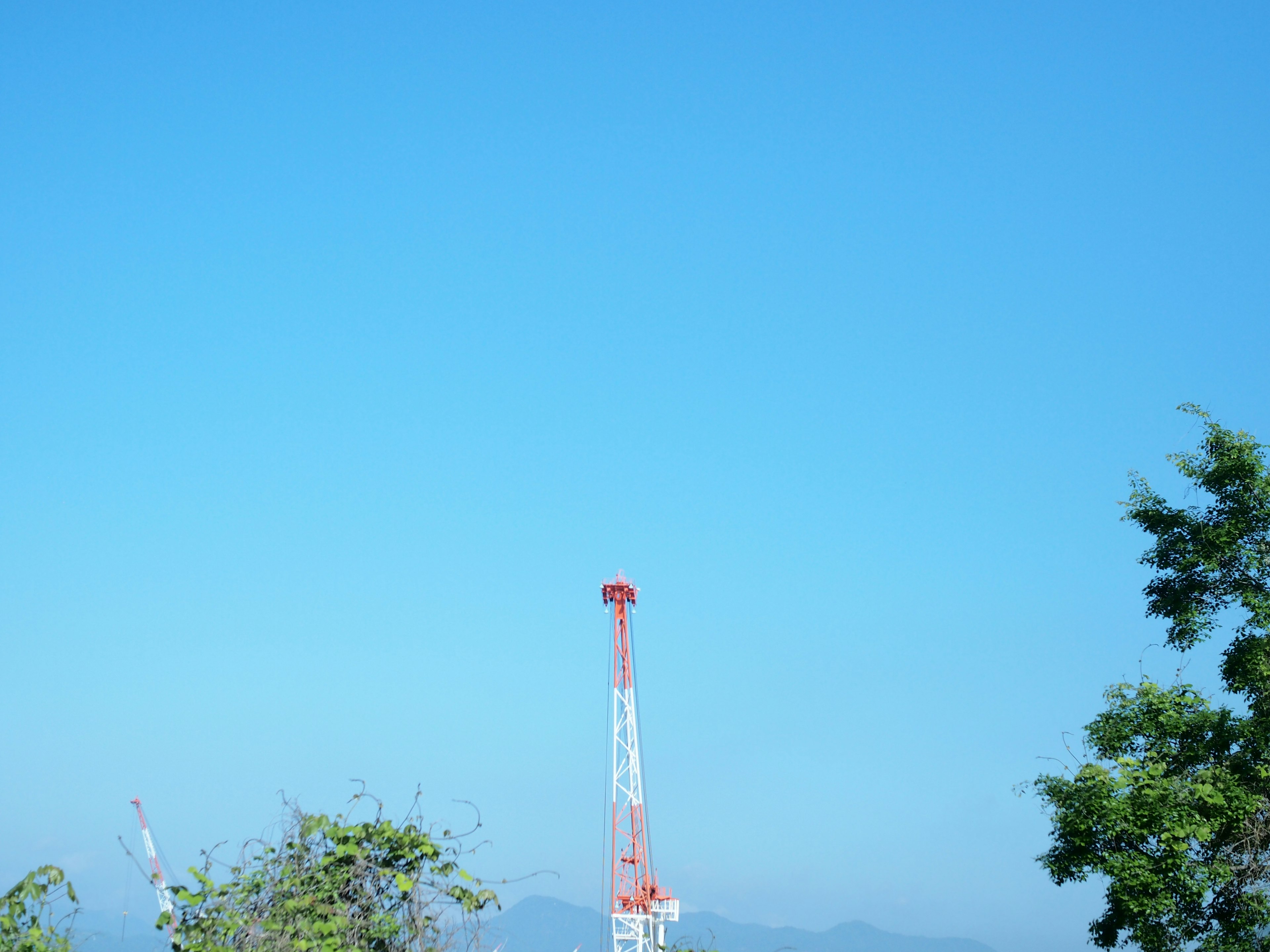 Red and white communication tower under a clear blue sky with surrounding greenery