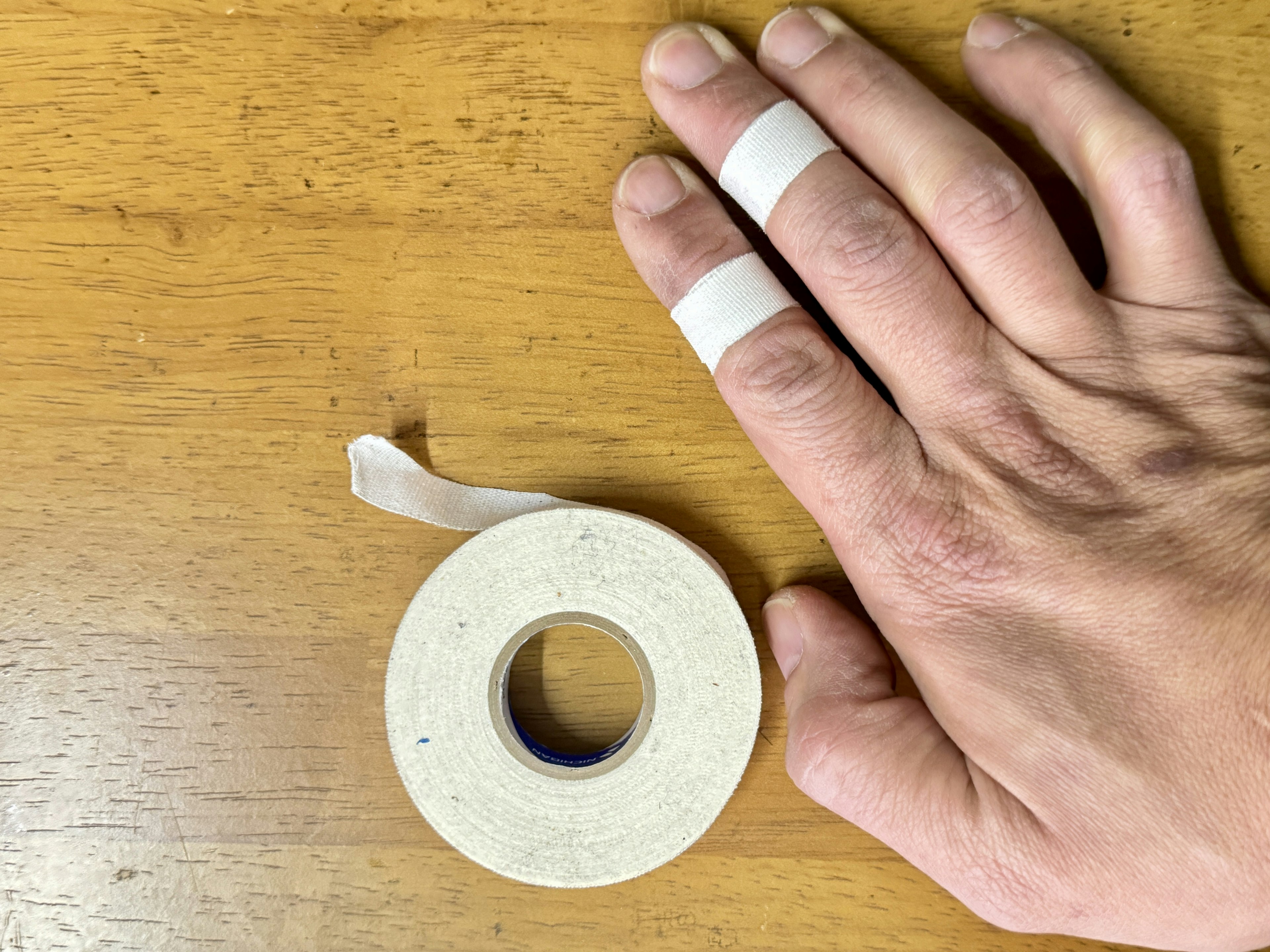 A hand with fingers wrapped in white tape next to a roll of white tape on a wooden surface