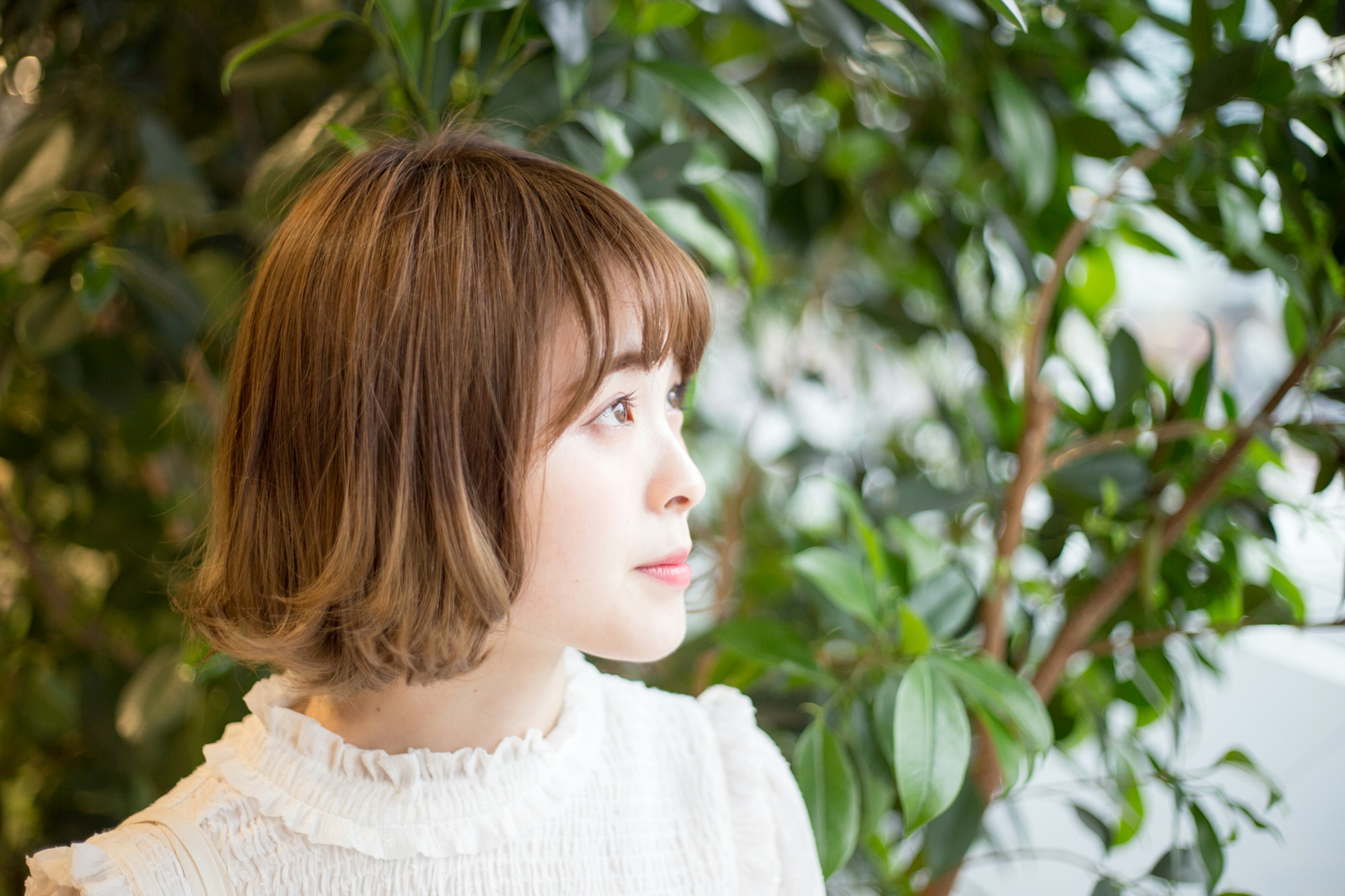 Perfil de una mujer sentada frente a plantas verdes, cabello castaño corto, vistiendo una blusa blanca