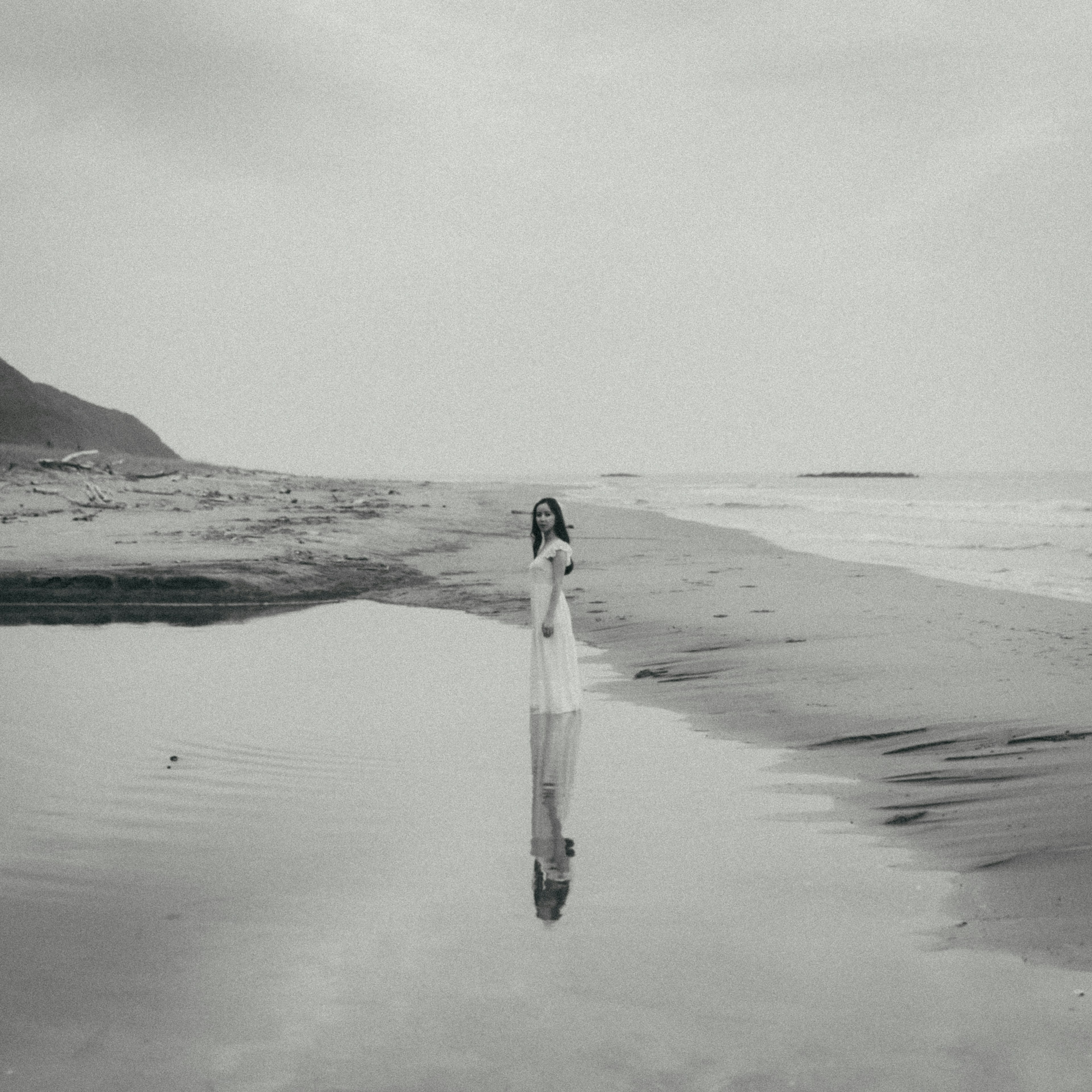 Schwarzweißfoto einer Frau in einem weißen Kleid, die in einer Pfütze am Strand steht