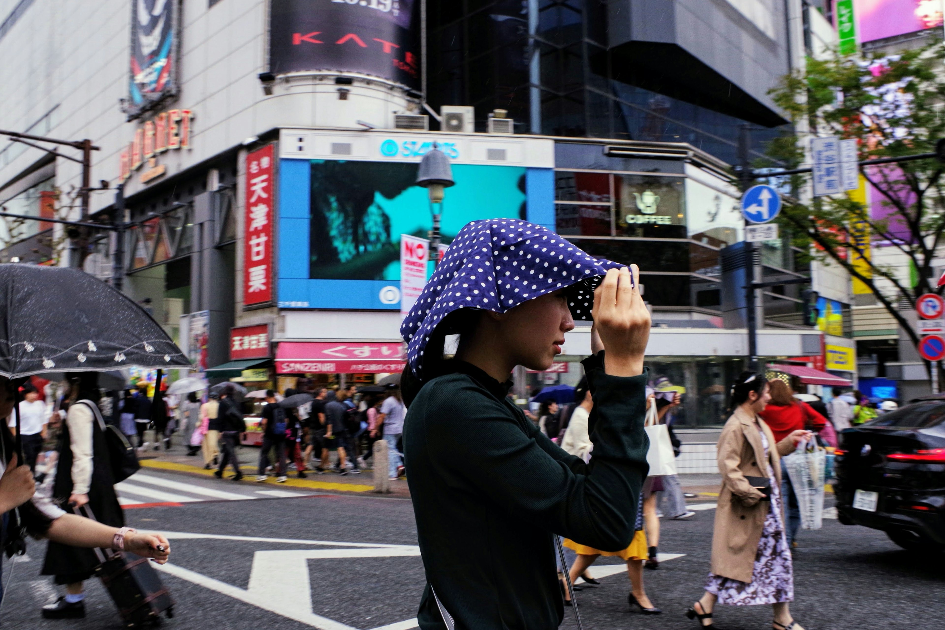 A busy urban street scene with a woman holding a hat wearing a polka-dot hood