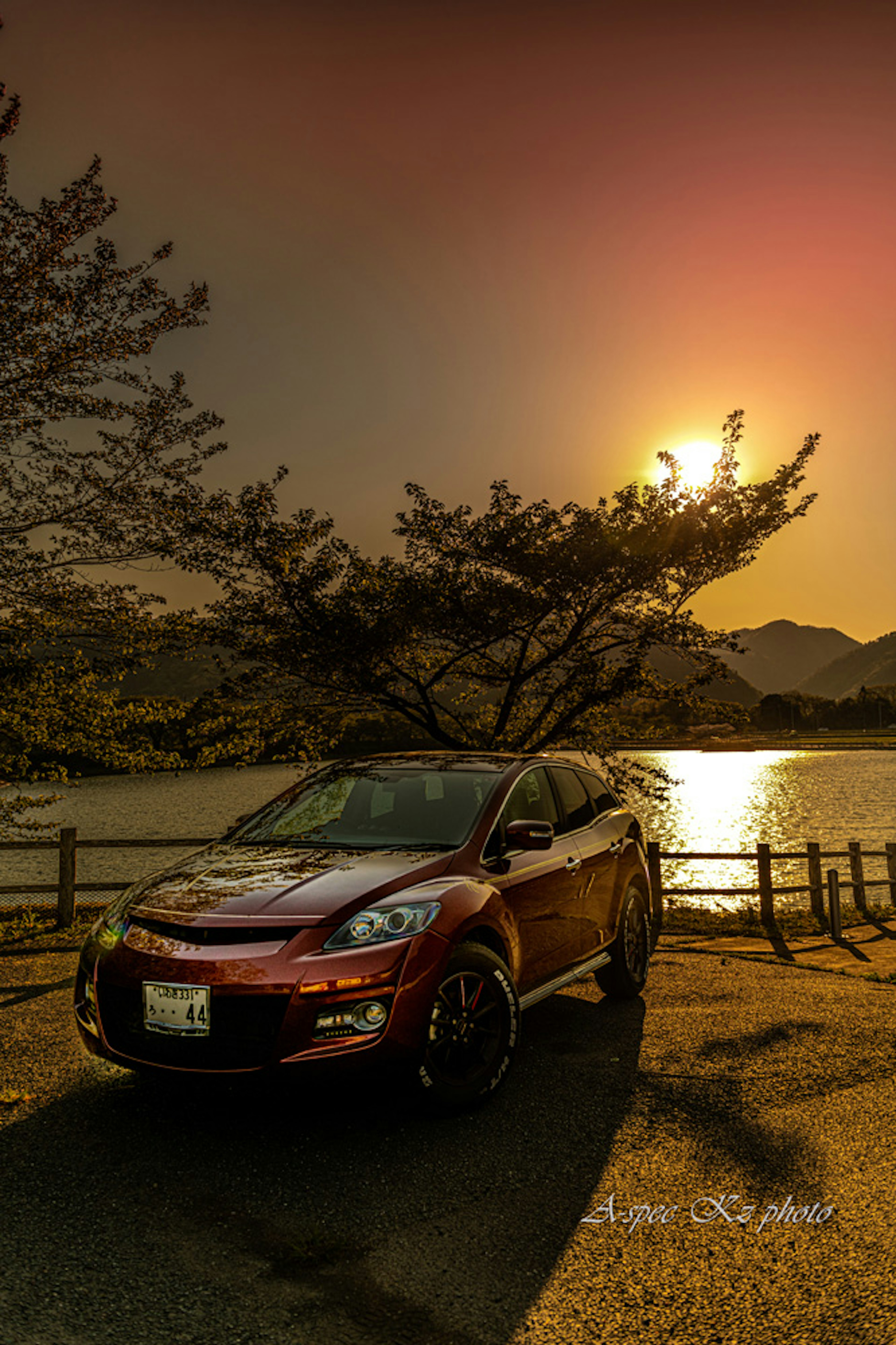 Red car parked near a lake with a sunset and trees in the background