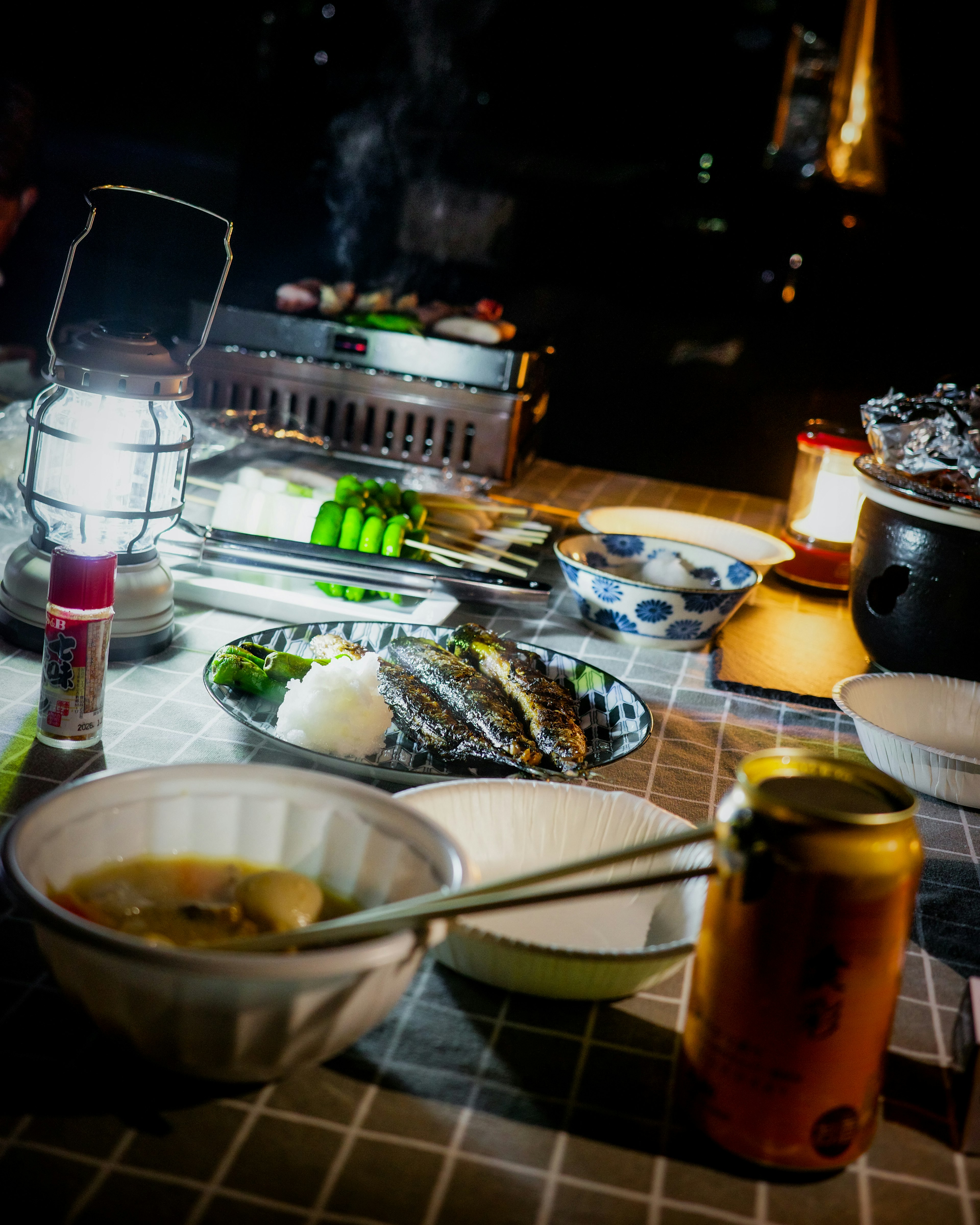 夜の屋外で食事を楽しむシーン　ランタンの明かりの下に料理と飲み物　焼き魚やご飯が並ぶ