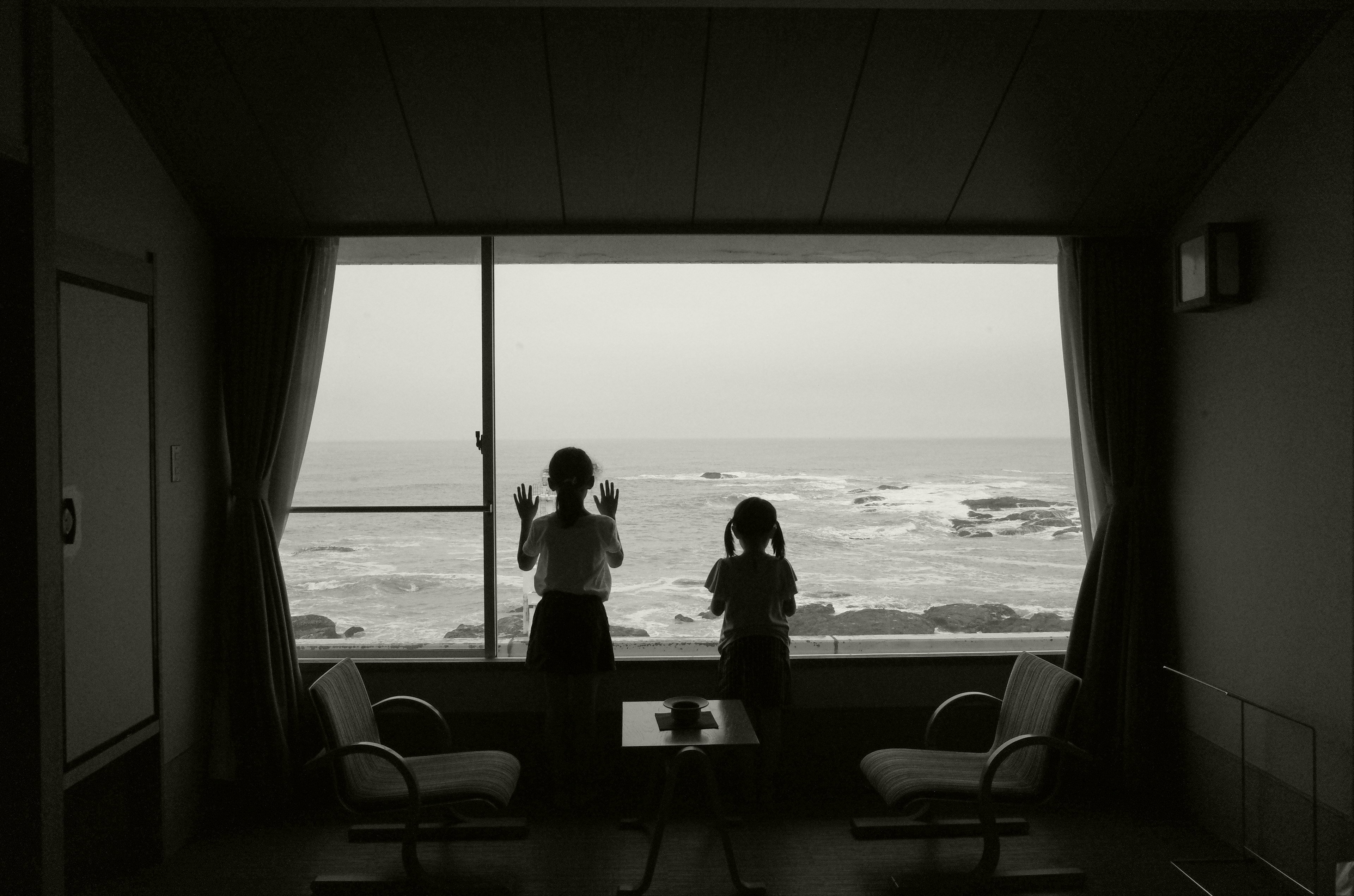 海を見つめる二人の子供と窓のある部屋のモノクロ写真