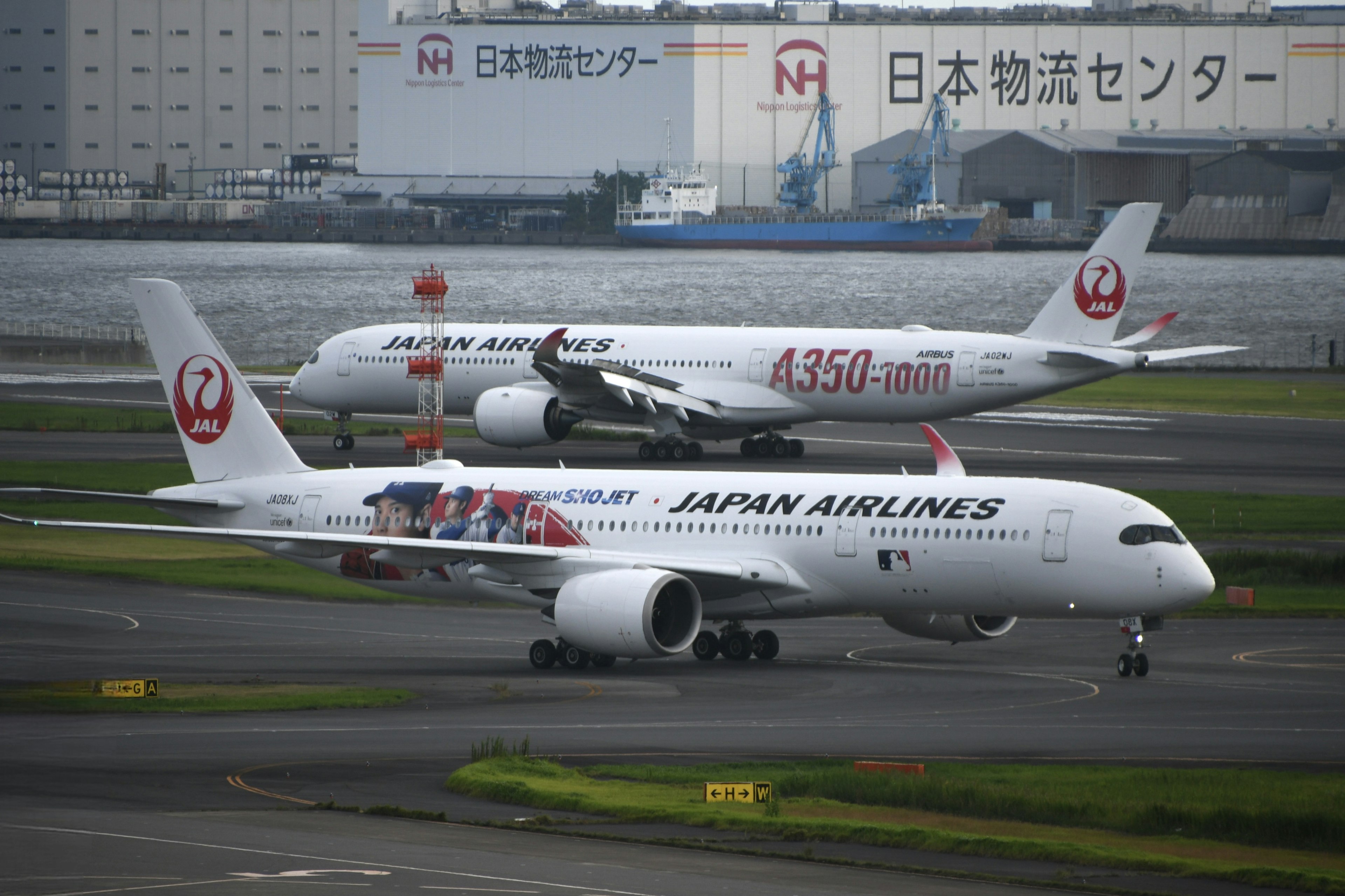 Máy bay chở khách Japan Airlines đang lăn trên đường băng với một máy bay khác và một kho hàng Nhật Bản ở phía sau