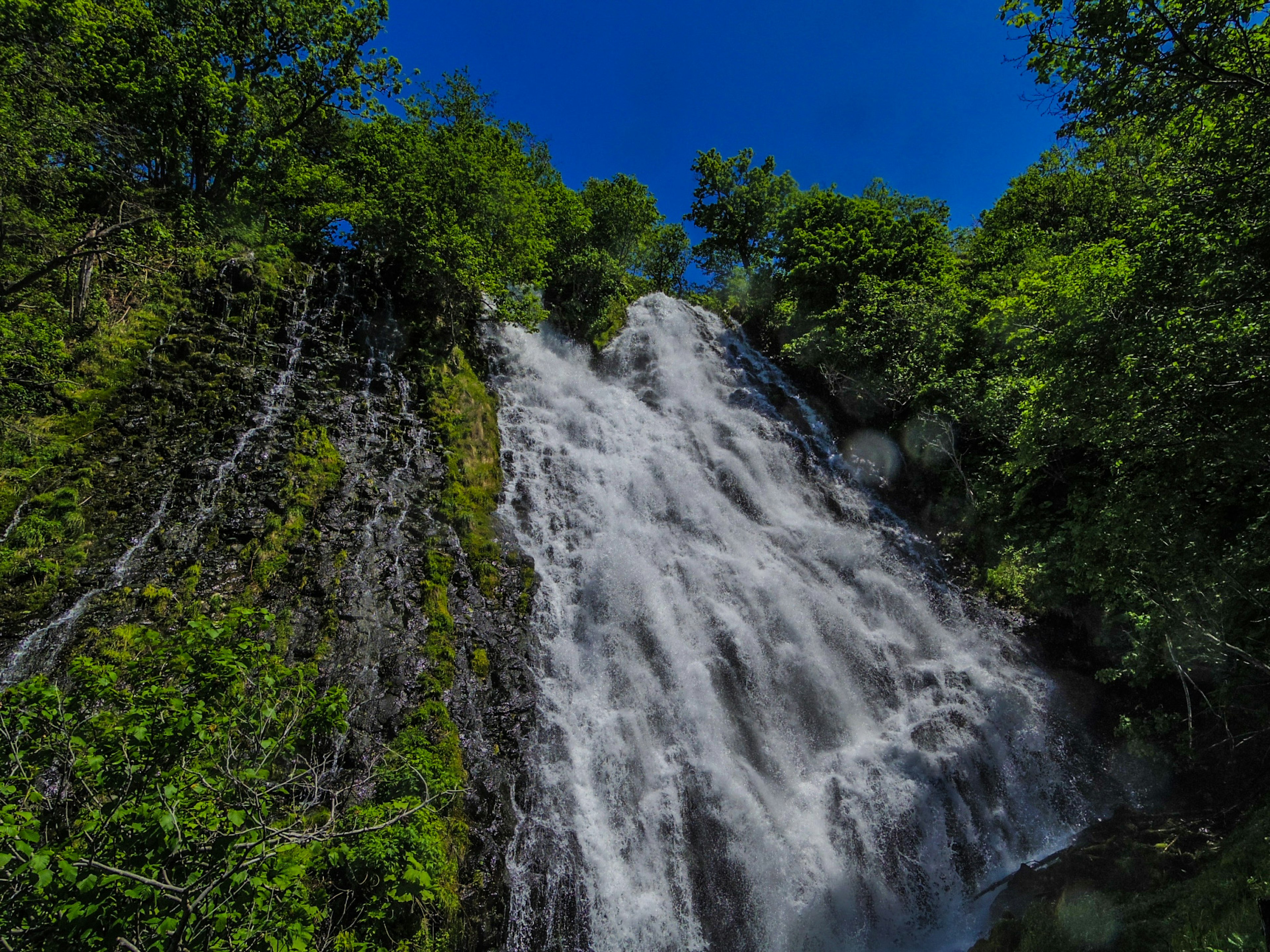 Air terjun yang indah mengalir di bawah langit biru dikelilingi oleh pepohonan hijau