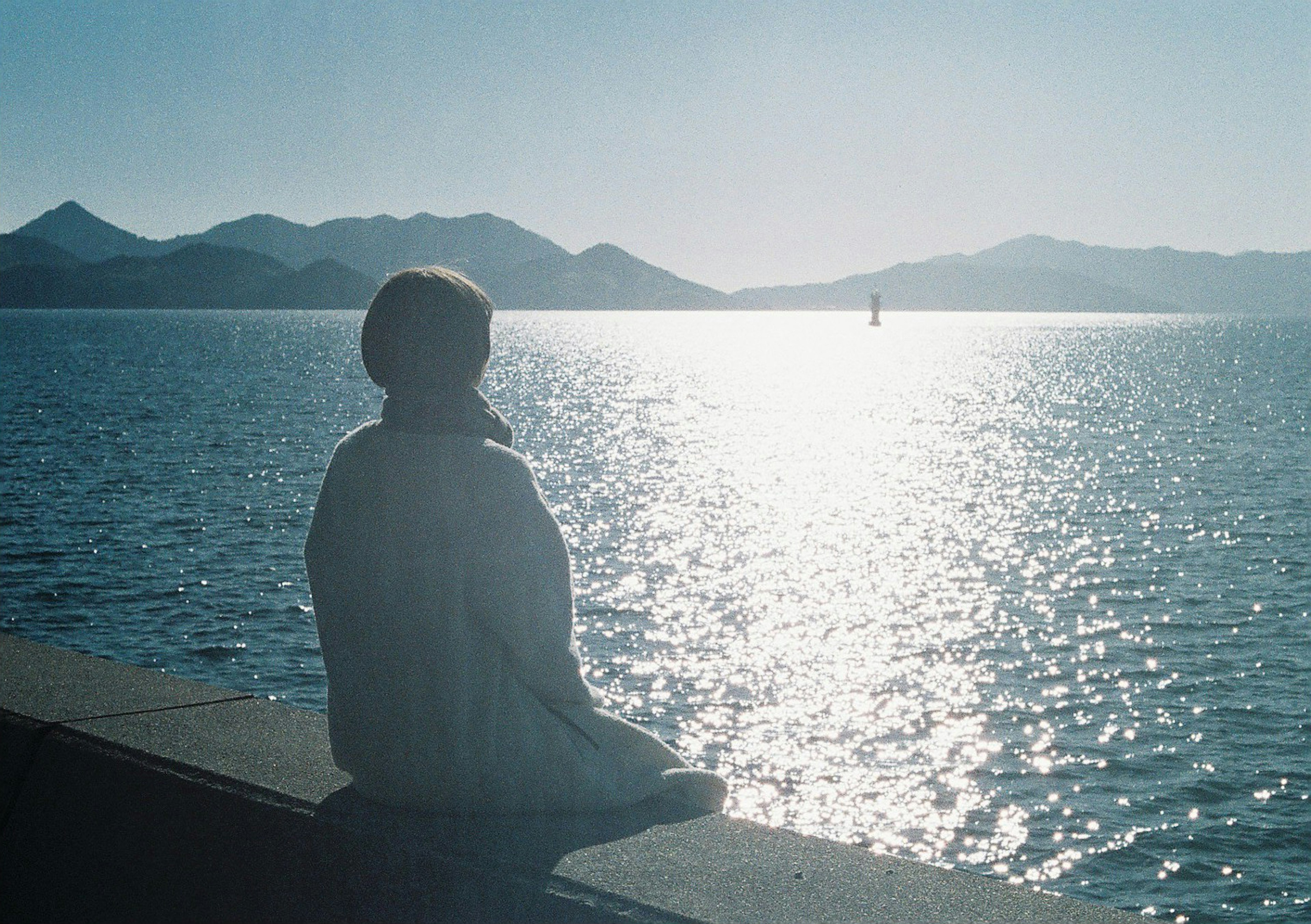 Silhouette einer Person, die an einem ruhigen See mit fernen Bergen sitzt