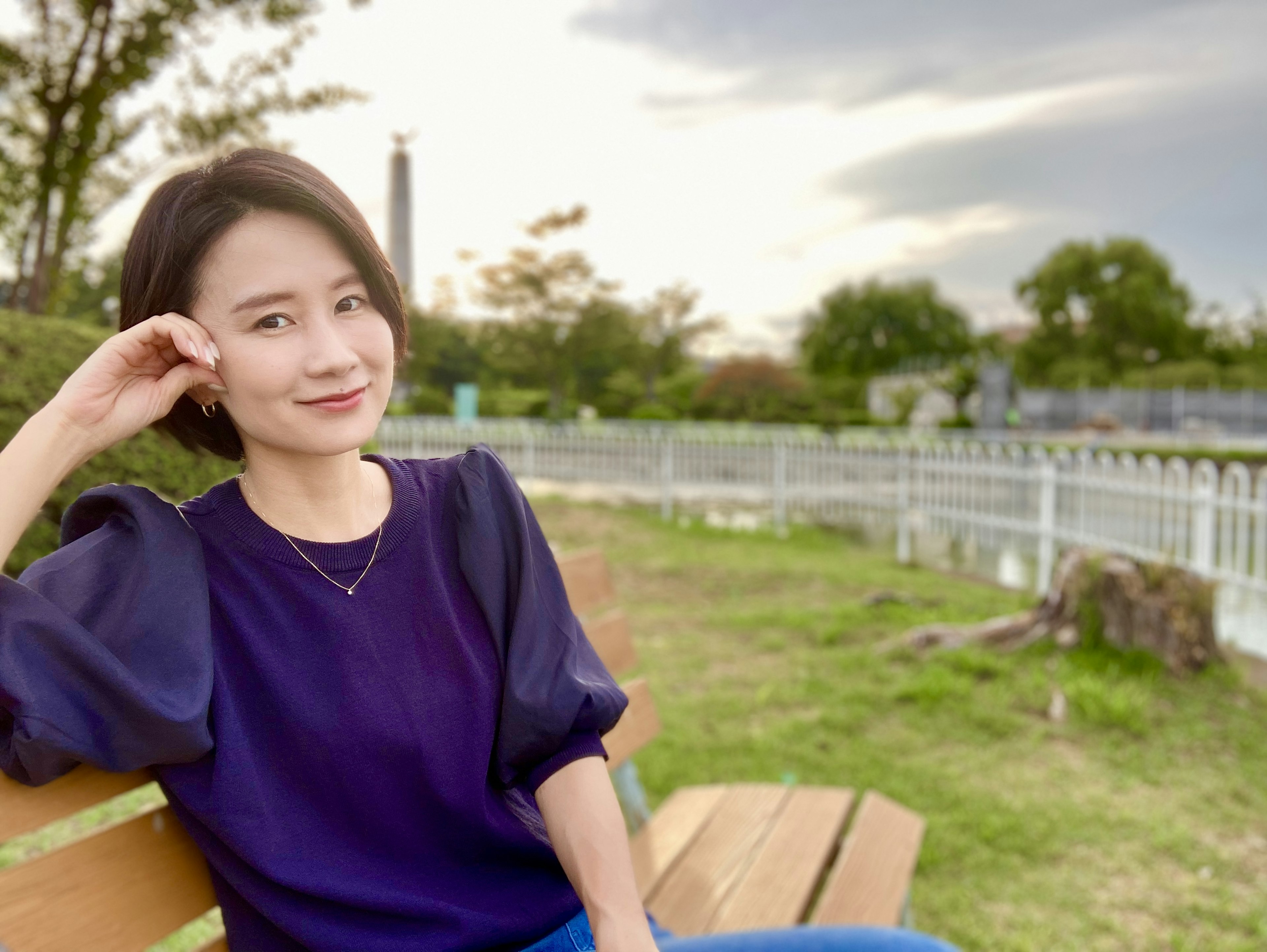 Woman sitting on a park bench wearing a purple top, calm expression, green trees and sky in the background