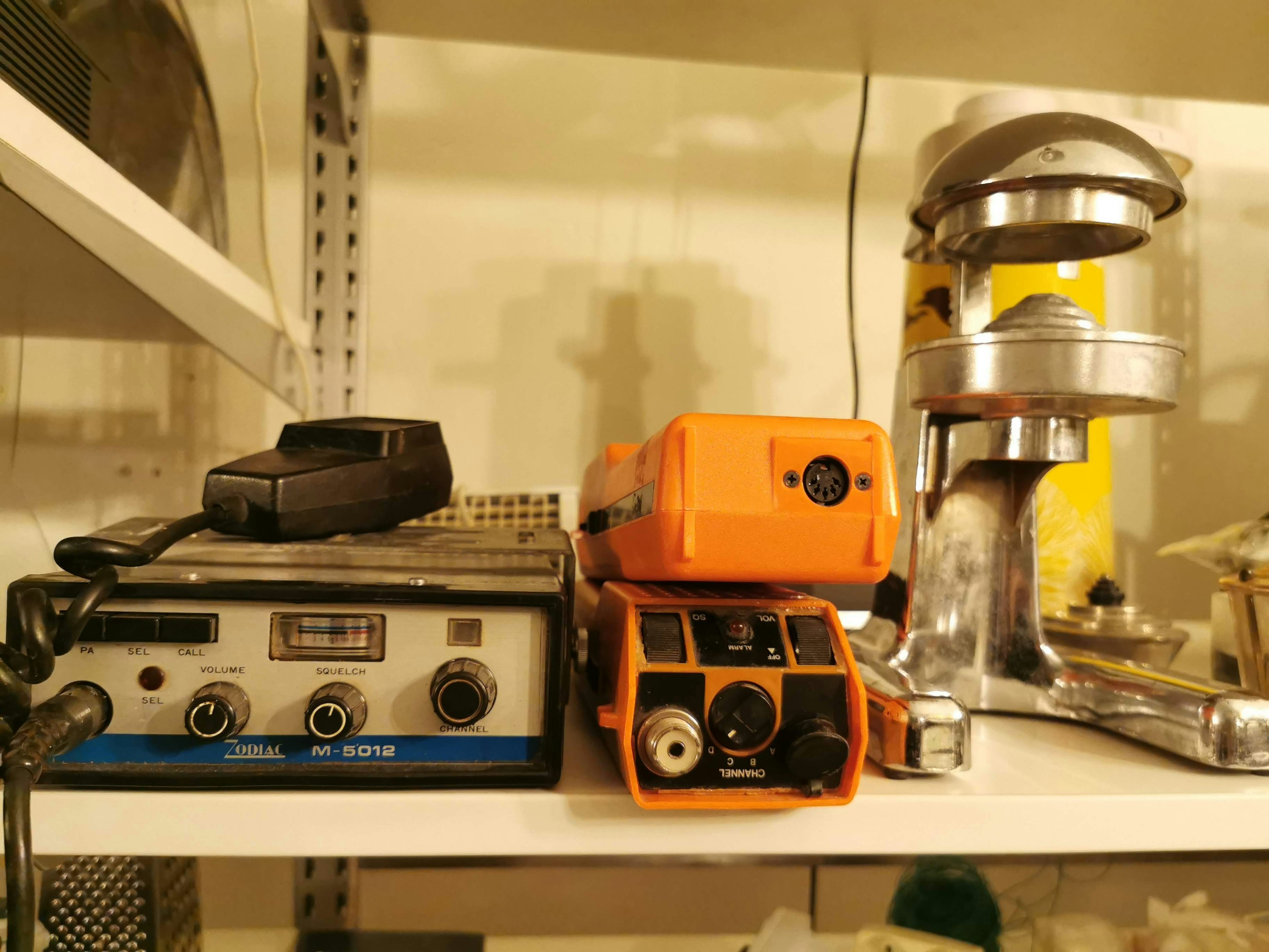 Image of old equipment on a shelf featuring a black radio device an orange device a metallic apparatus and a yellow container