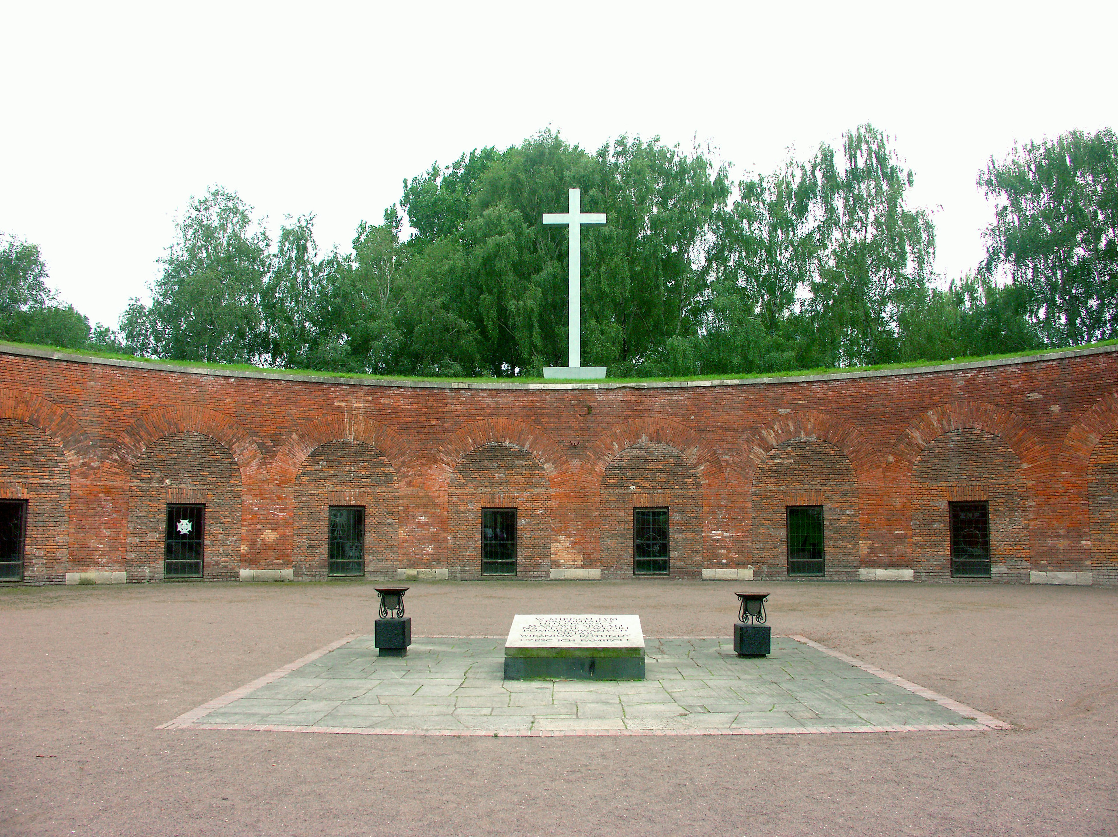 Weiße Kreuz in einem Innenhof, umgeben von roten Backsteinmauern und einem Steinaltar