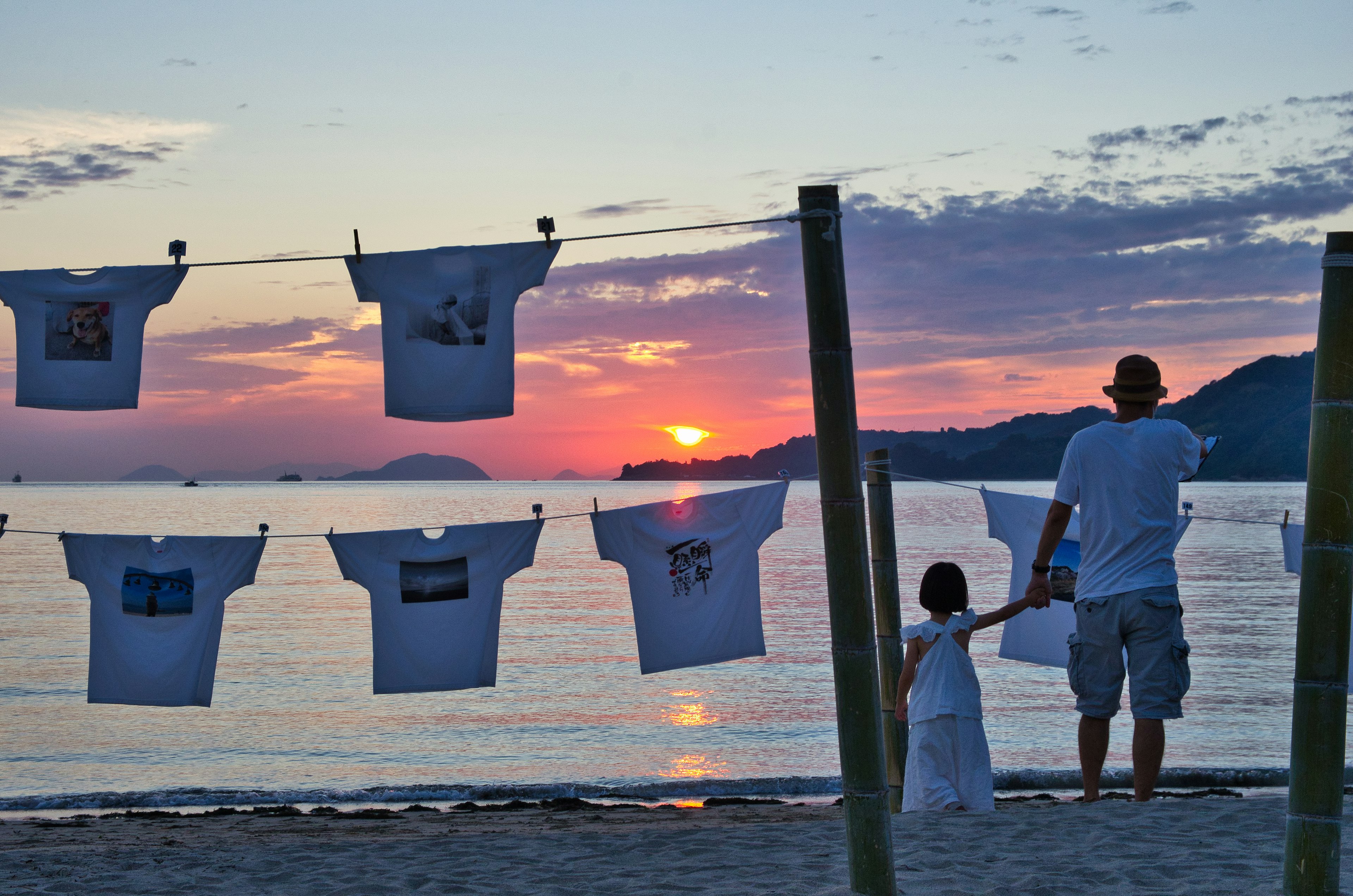 夕日を背にした父と子供が海辺で立っている Tシャツが干されている