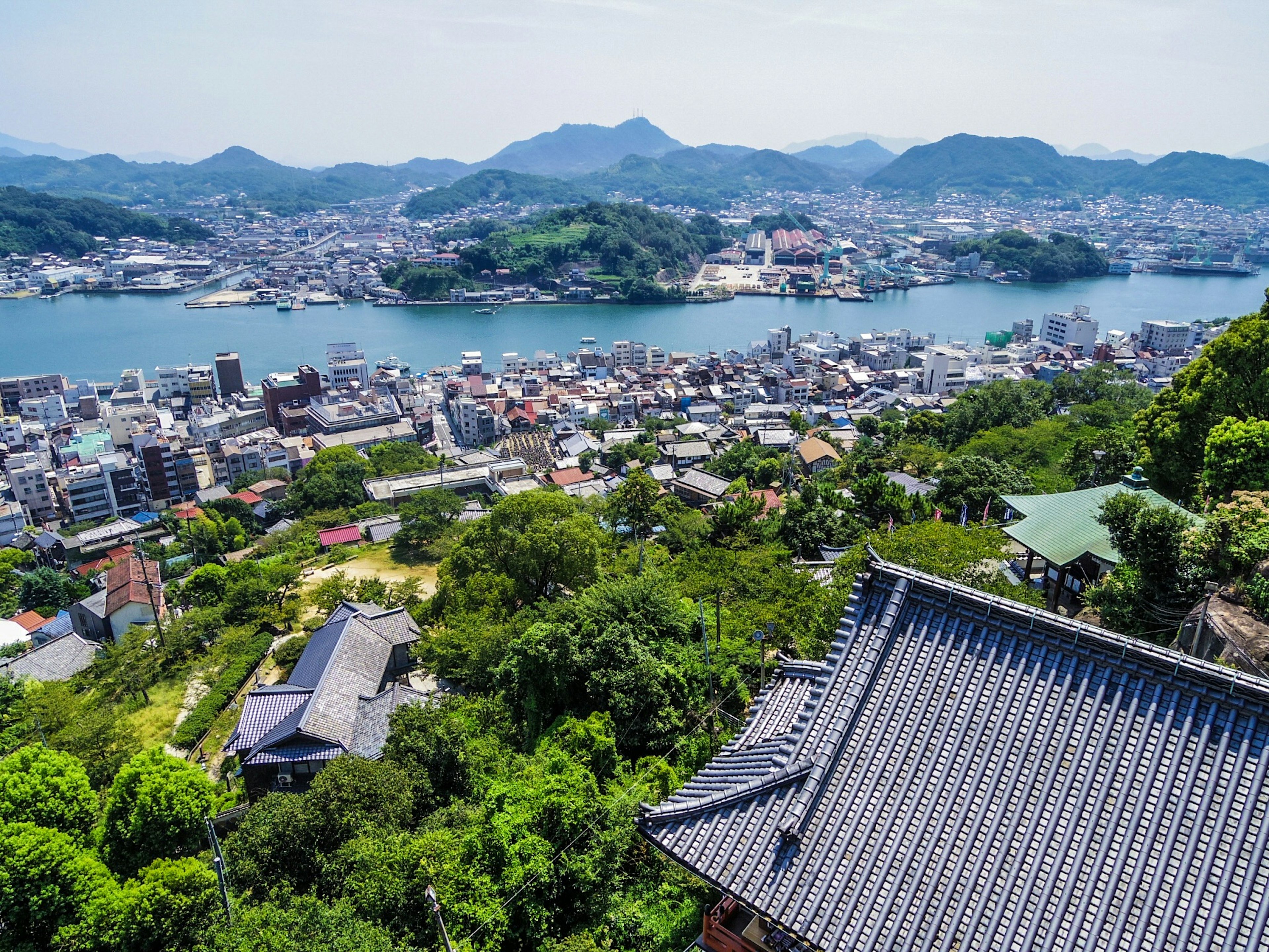 Cityscape featuring lush mountains and sea backdrop with traditional Japanese building roof