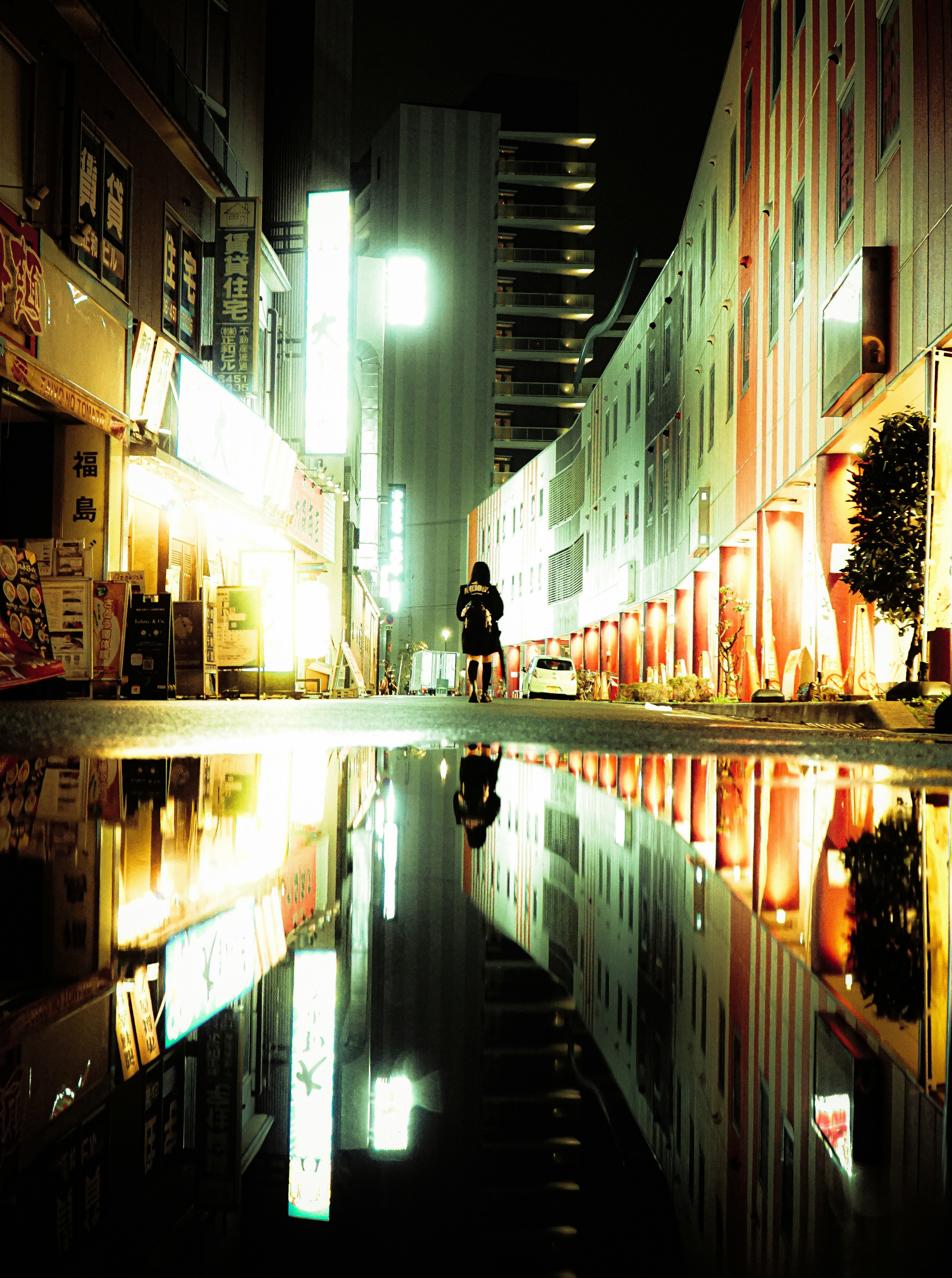 Esquina de calle nocturna con luces de neón reflejadas en un charco y la silueta de una persona