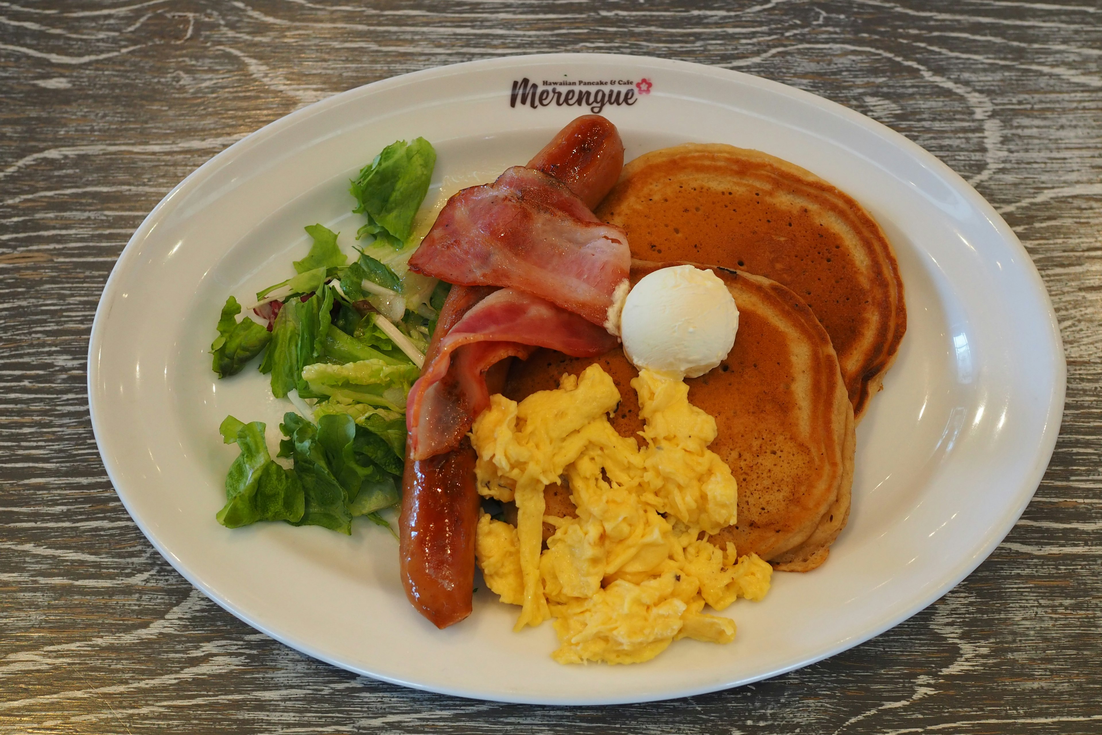 A plate with pancakes, bacon, sausage, scrambled eggs, and salad