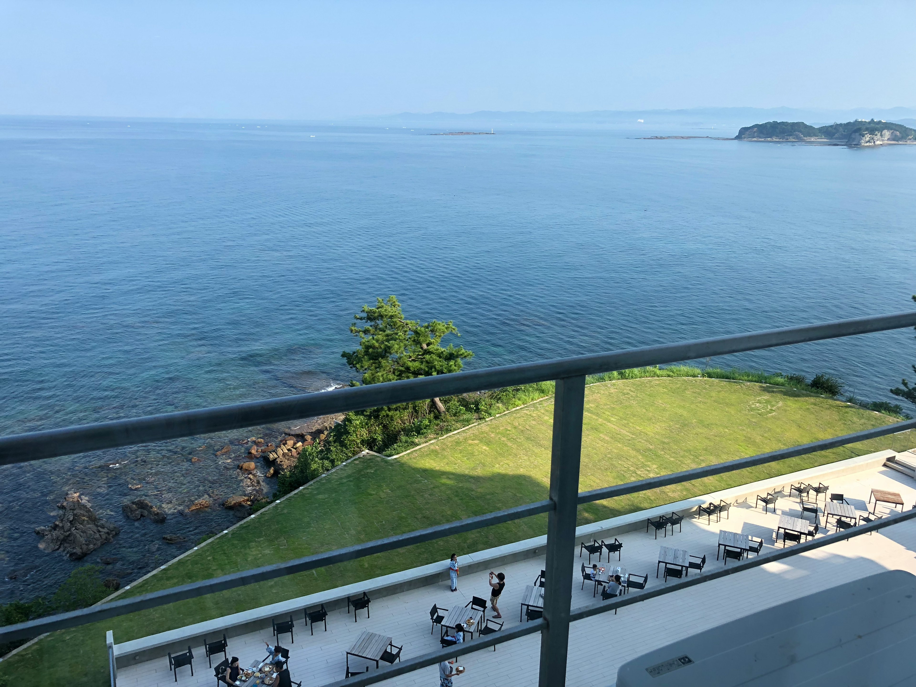 青い海と明るい空を背景にしたバルコニーの景色　海岸に沿った芝生と日光浴用の椅子が見える