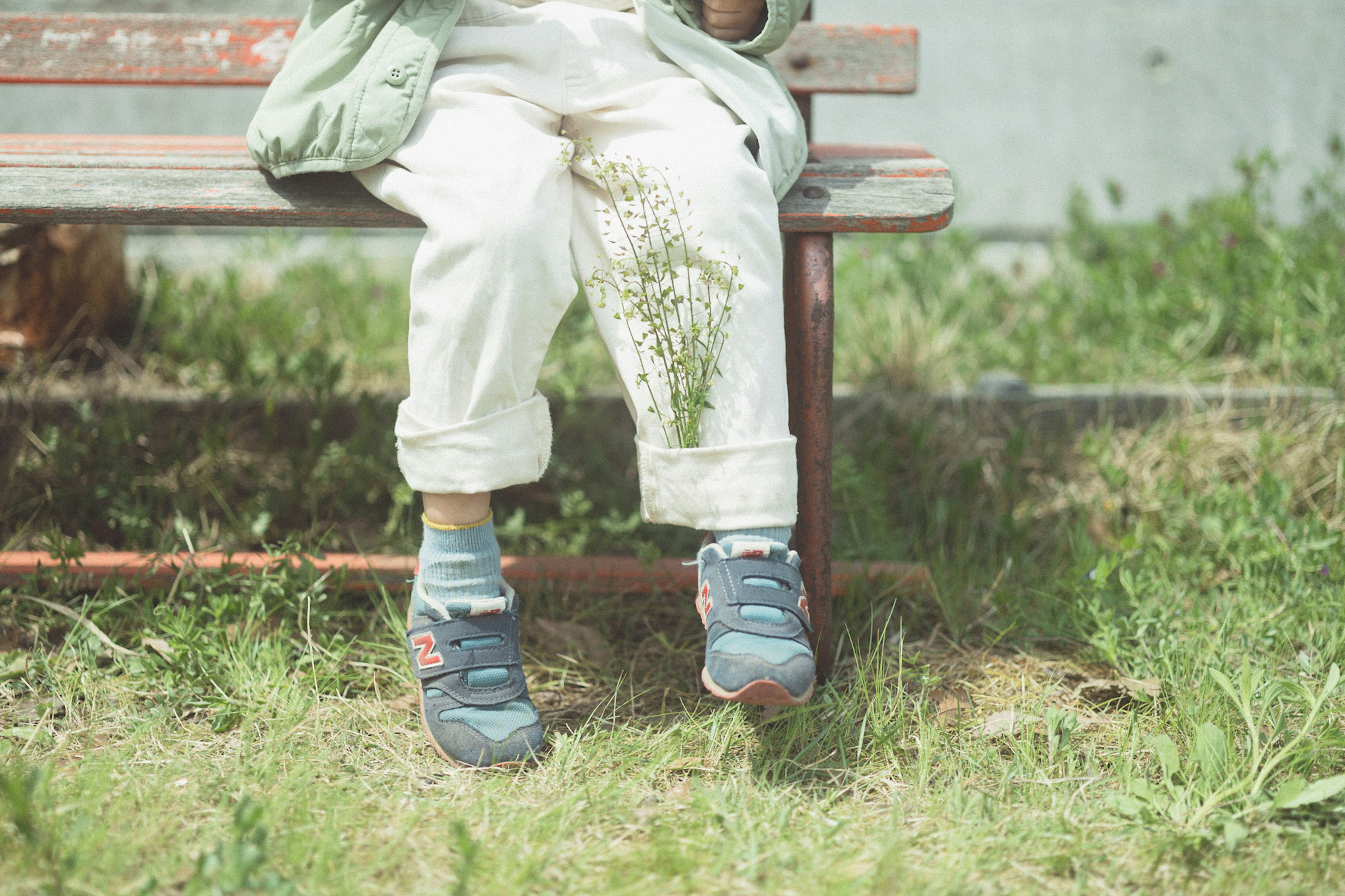 Enfant assis sur un banc de parc avec un pantalon blanc sale et des chaussures bleues entouré d'herbe verte