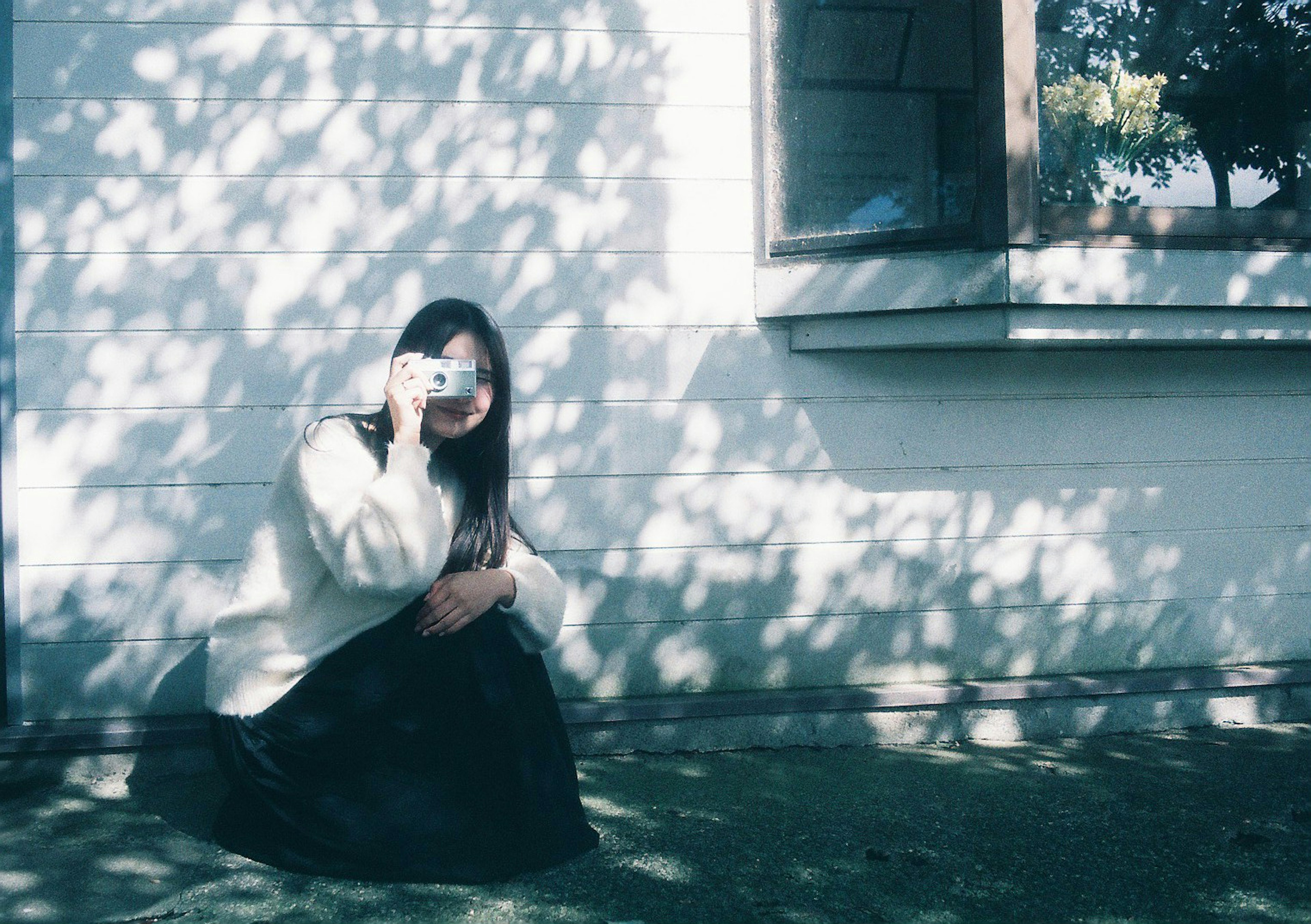 Una donna con una macchina fotografica seduta vicino a una finestra con ombre su un muro bianco